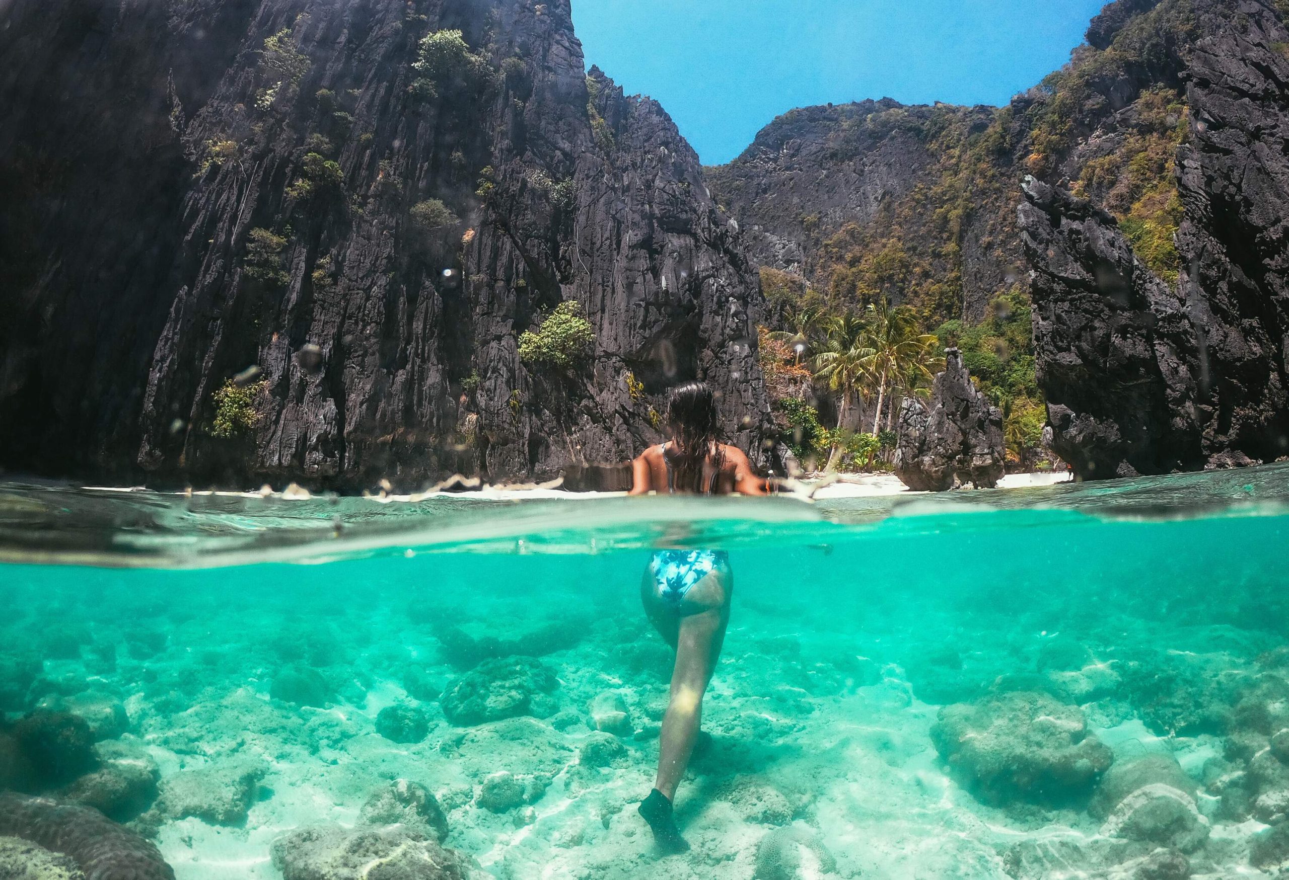 A woman in swimwear submerged in the crystal-clear waters of a beach, surrounded by towering blackish rocks.