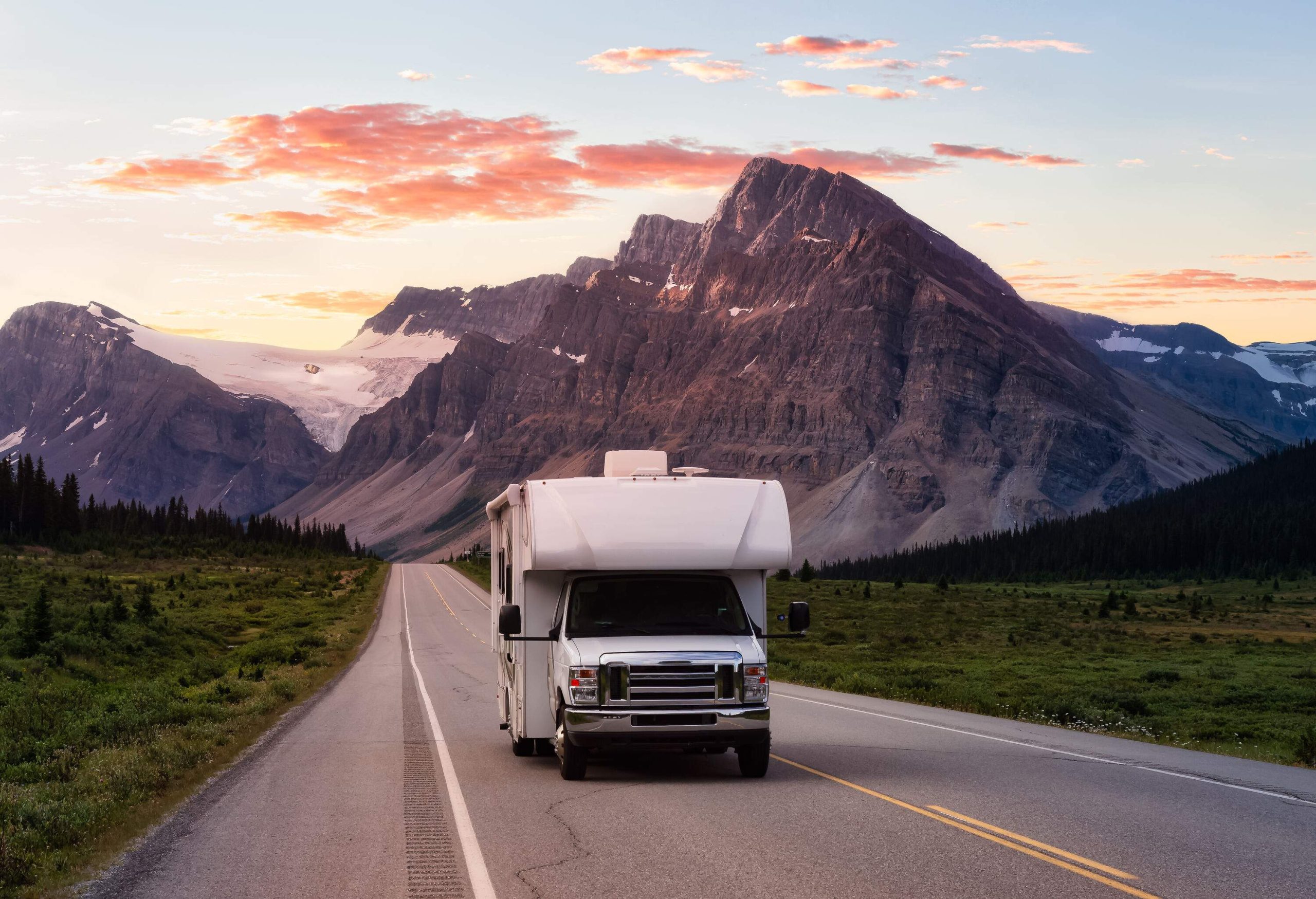 White camper van driving on an empty road at sunrise with views of snow-covered mountains.