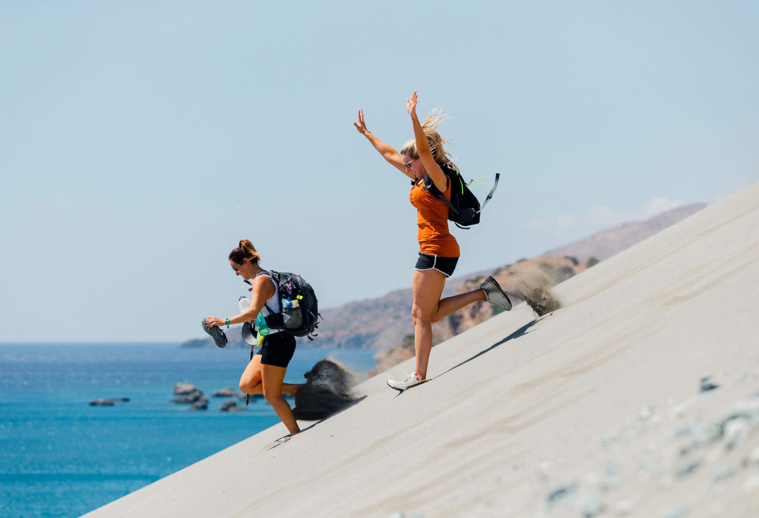 Two women with bags sprint down a sloping sand dune next to the sea.