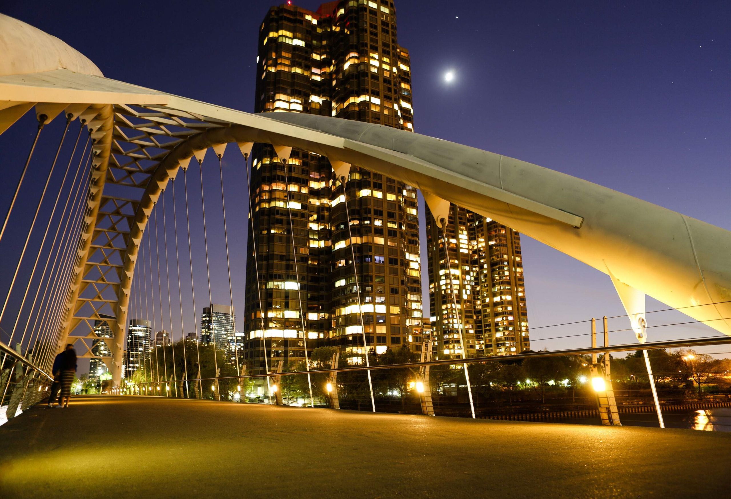 A couple on an illuminated through-arch bridge on the far side of a tall building.