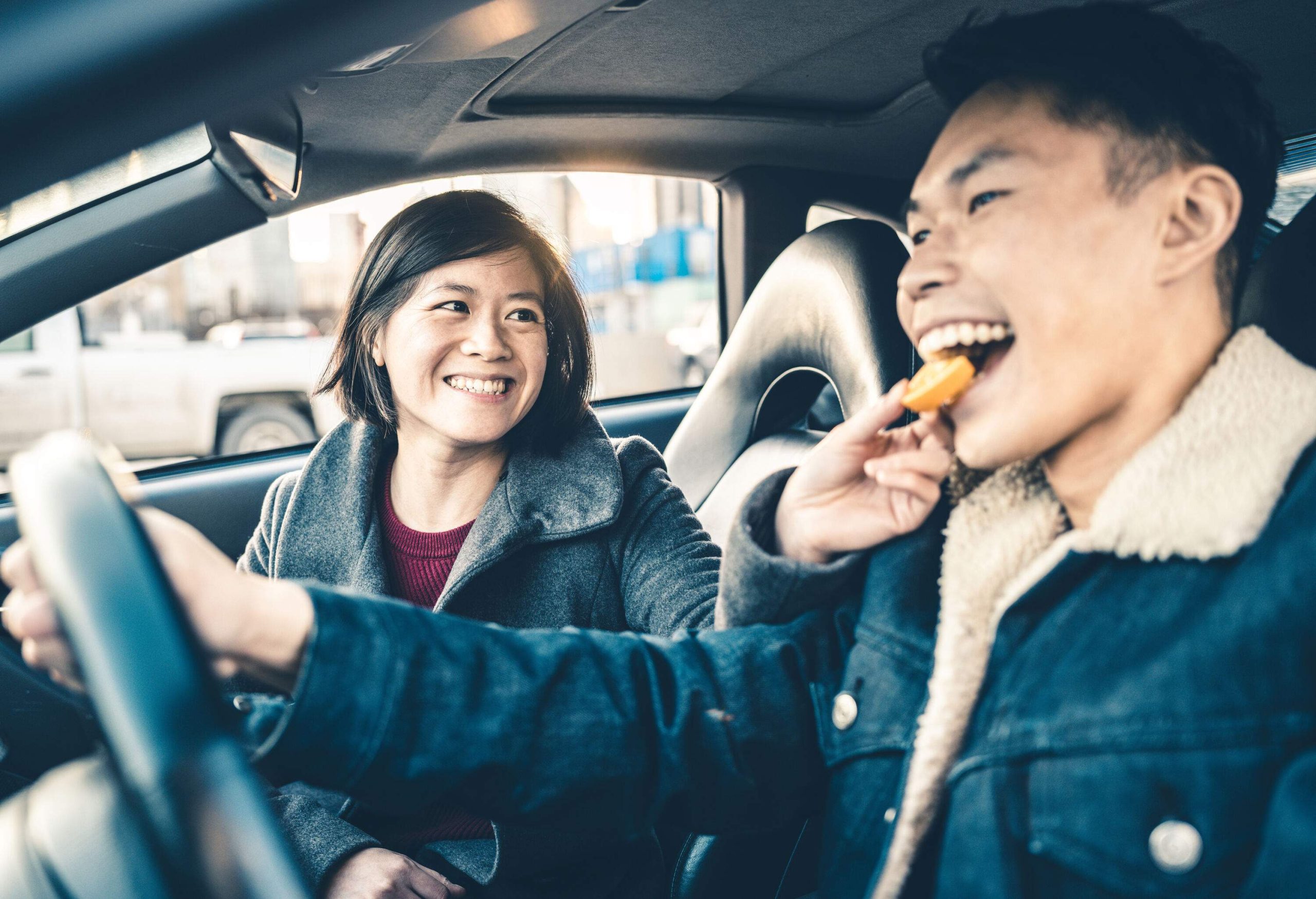 A man drives a car while a woman sitting on the passenger's seat feeds him a cookie.
