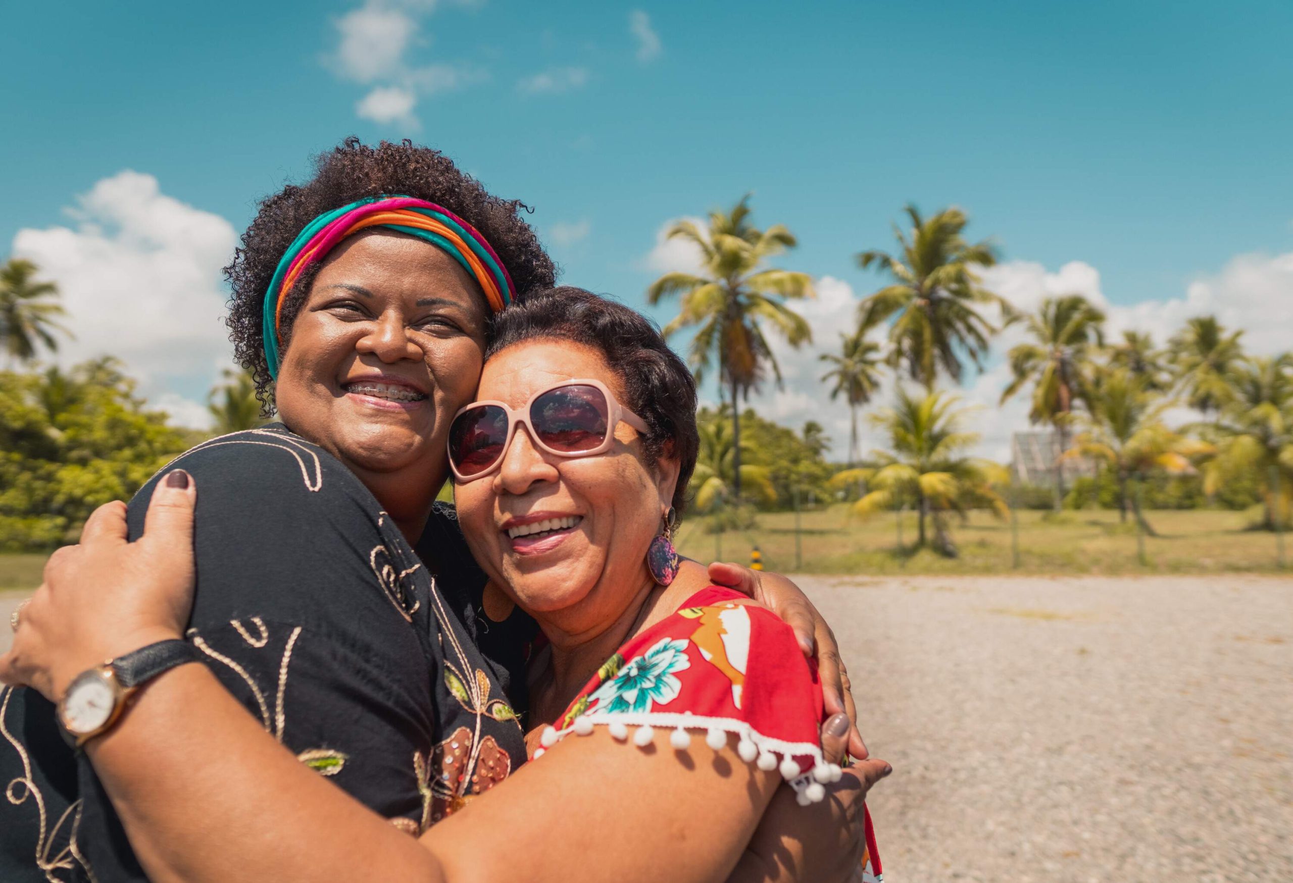 Two friends share laughter and a warm embrace on a sun-soaked beach, surrounded by nature, as one sports a stylish pair of sunglasses.