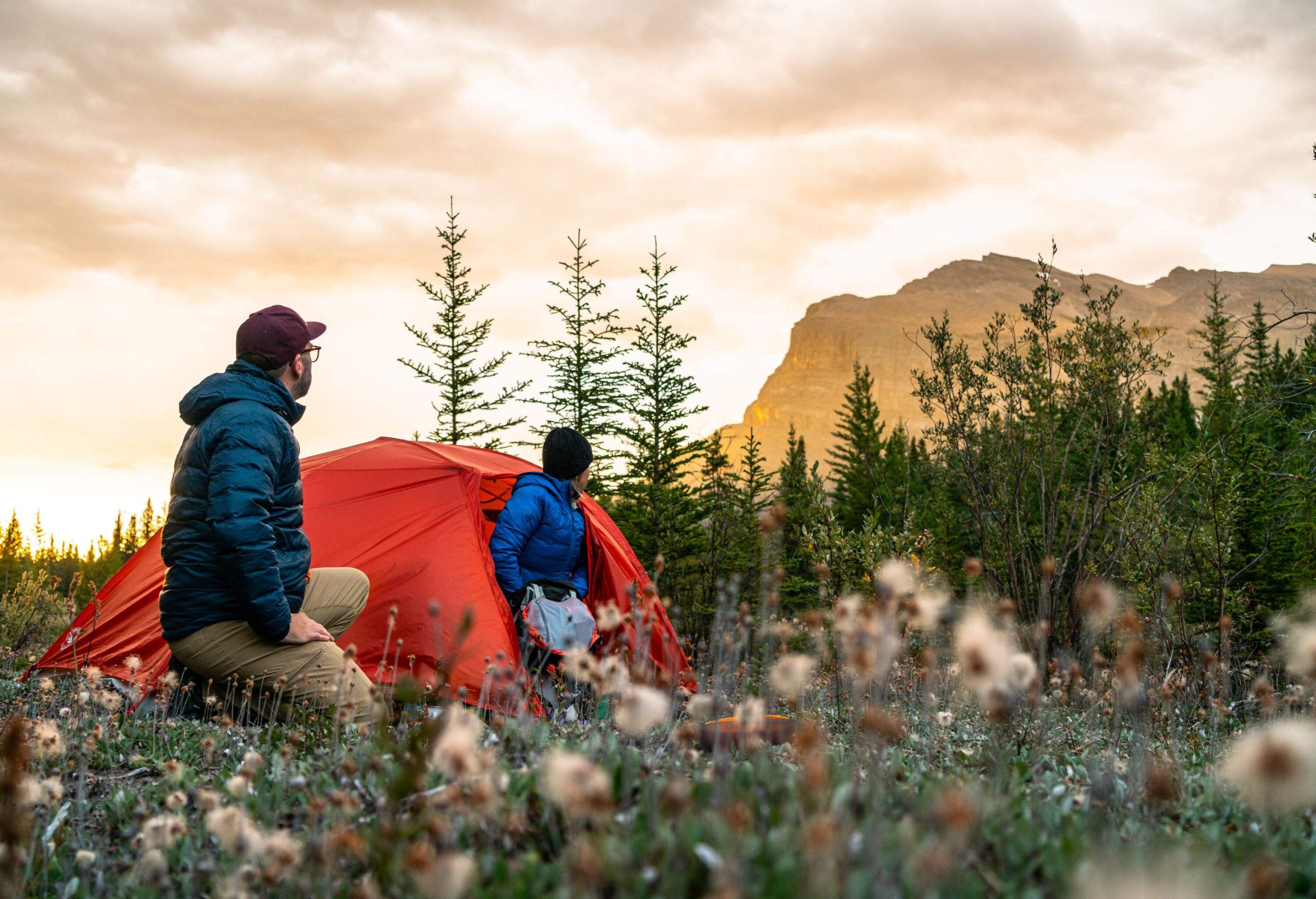 Two campers in puffer jackets look at a distant mountain from their orange tent.