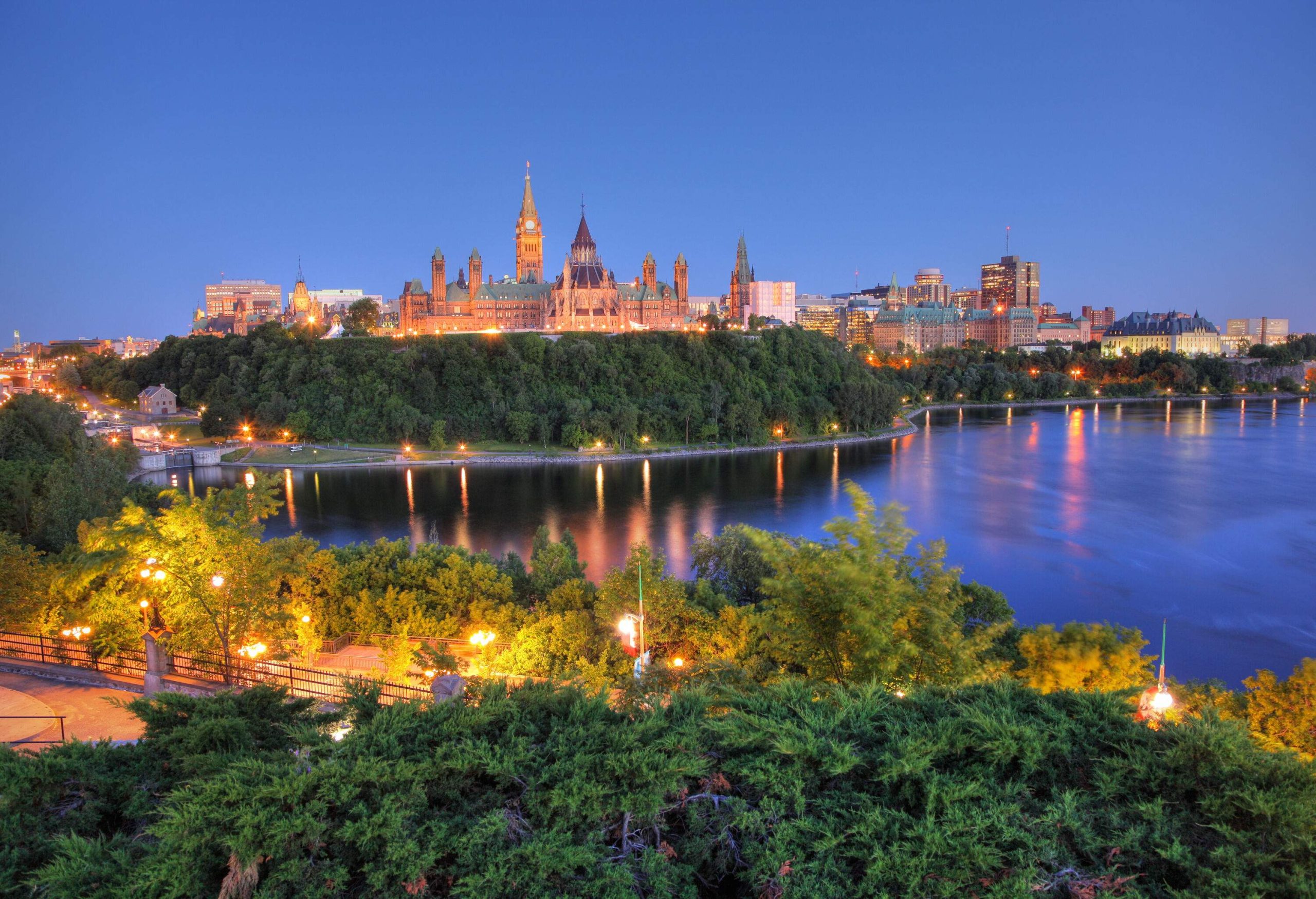 Breathtaking view of a serene river winding through a lush landscape, with a brightly lit city featuring modern buildings and a majestic Gothic Revival government building resembling a palace.