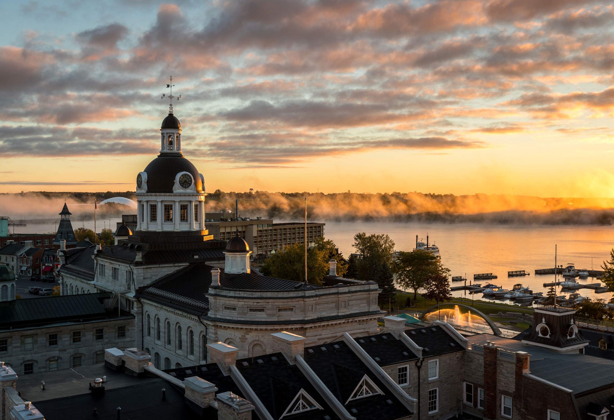 A building with a pillared dome roof and cupola overlooks a foggy river illuminated by the warm lights of a sunrise.