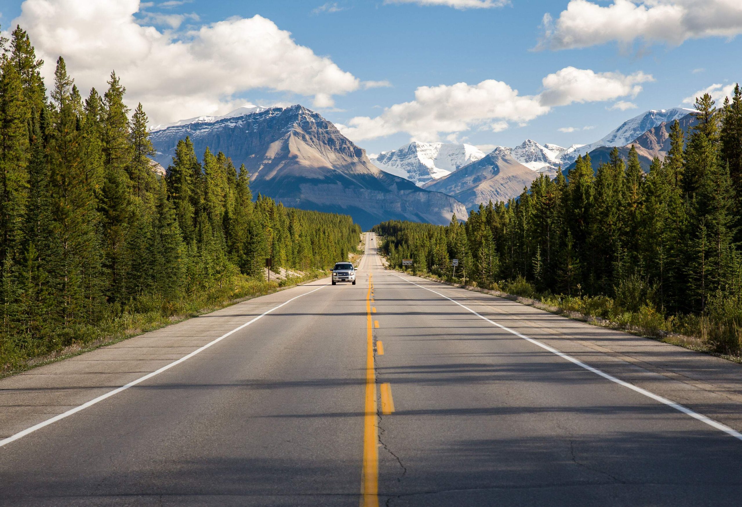 A grey car traversing the long road between lush green trees with stunning sharp-pointed rock mountains behind.