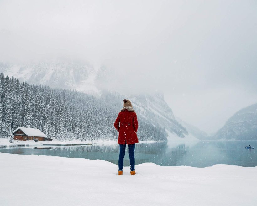 A woman in winter clothes standing in front of a lake, with a cabin and a misty forest in the background.