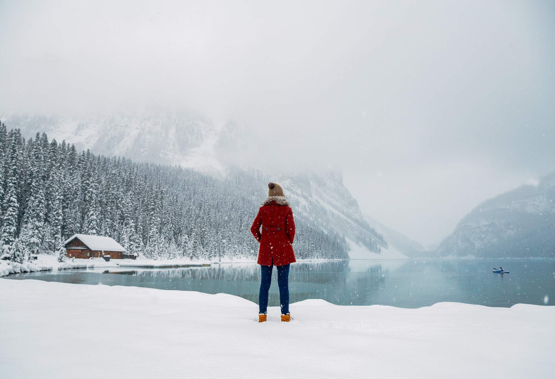 A woman in winter clothes standing in front of a lake, with a cabin and a misty forest in the background.