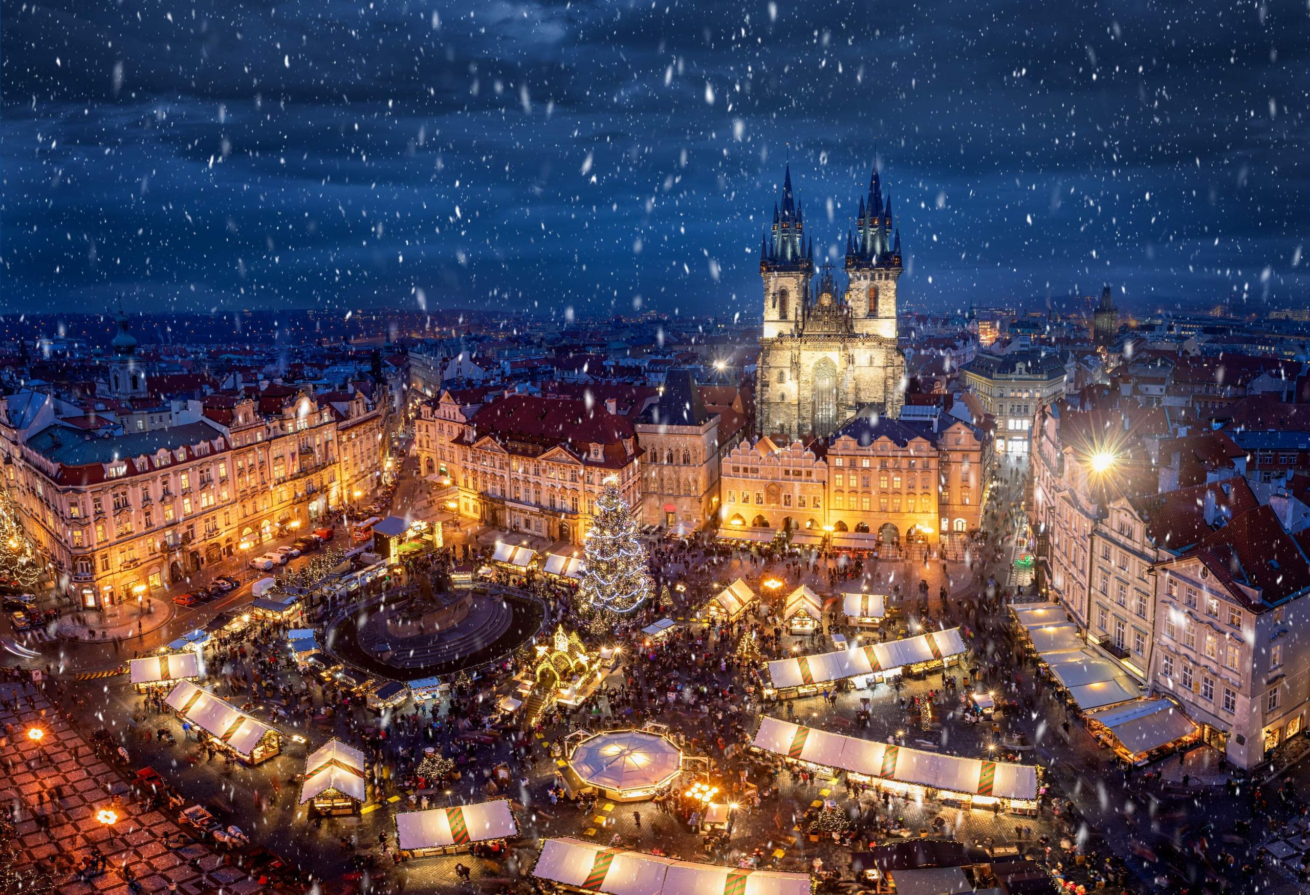 Snowflakes fall into the brightly lit, bustling square framed by buildings on a scenic festive Christmas season.