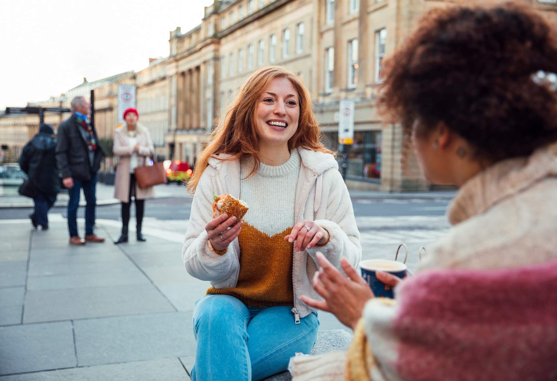 A young mixed-race woman and a young caucasian redhead woman sitting down together on a bench in the city and enjoying a bite to eat.