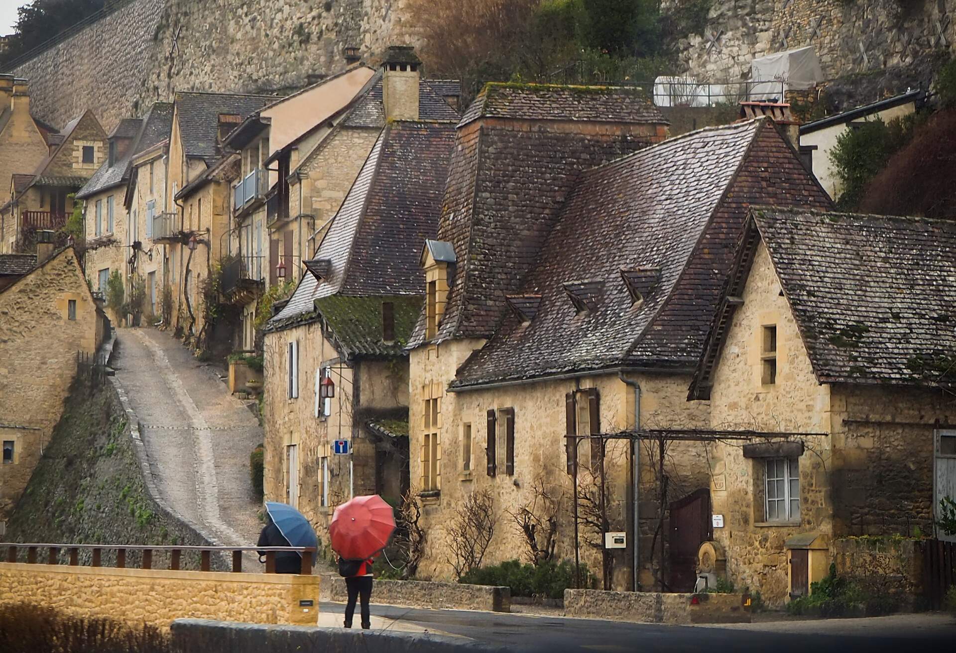 People with umbrellas stand outside the dirty-looking classic hillside stone houses.