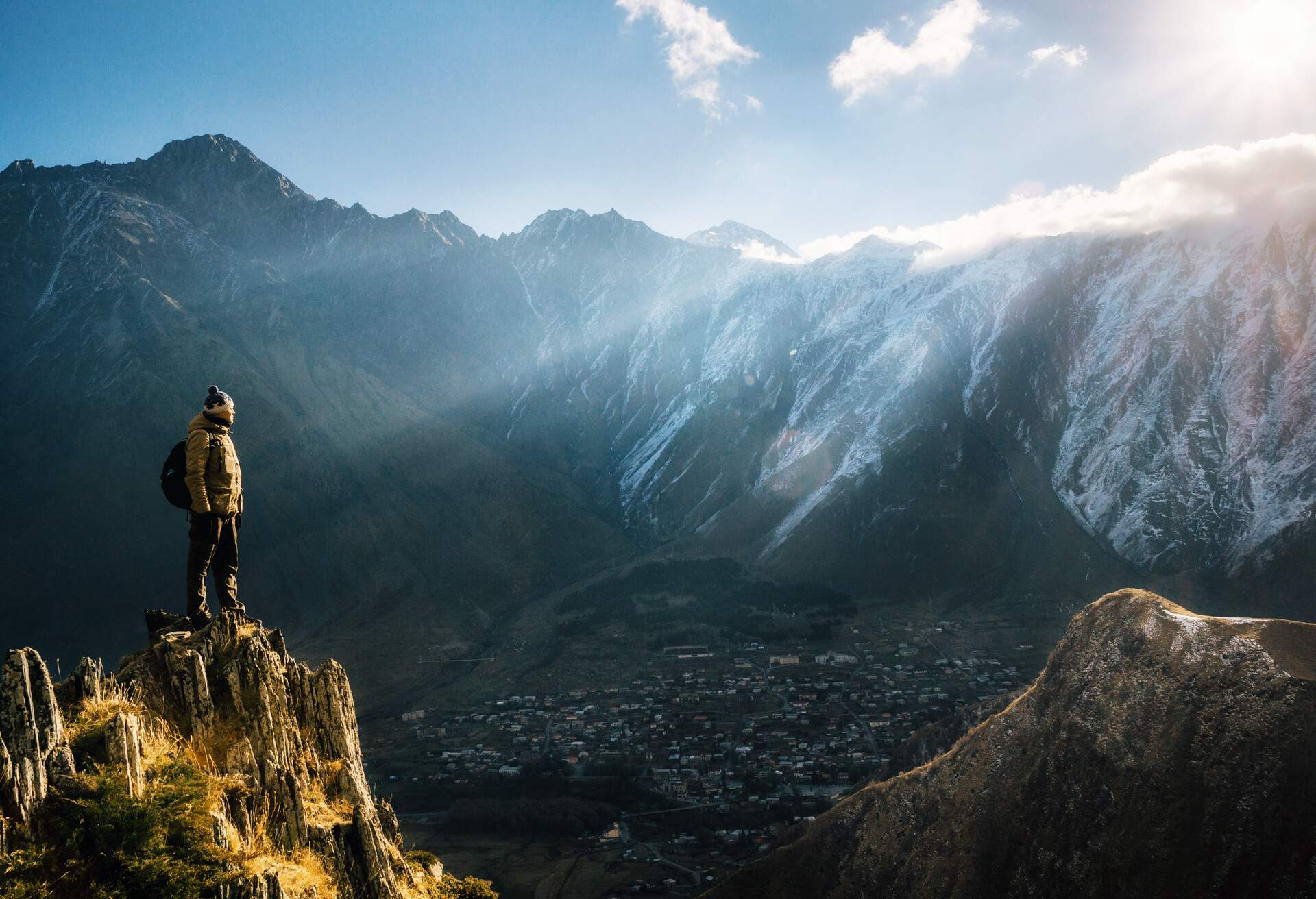 Young tourist in bright hat, black trousers with a backpack stands on cliff's edge and looking at the misty mountain village and glacier at sunrise, Stepantsminda, Georgia