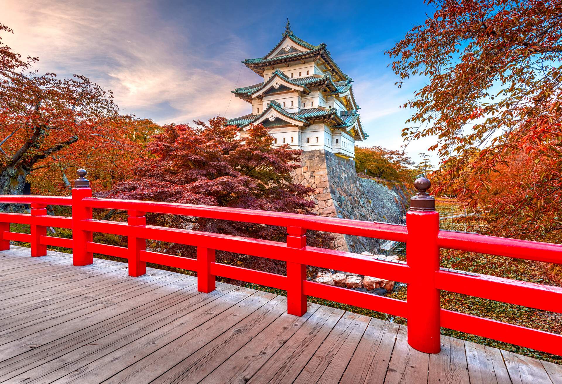 A wooden bridge with red railings over a moat backdropped by a three-story Japanese castle. 