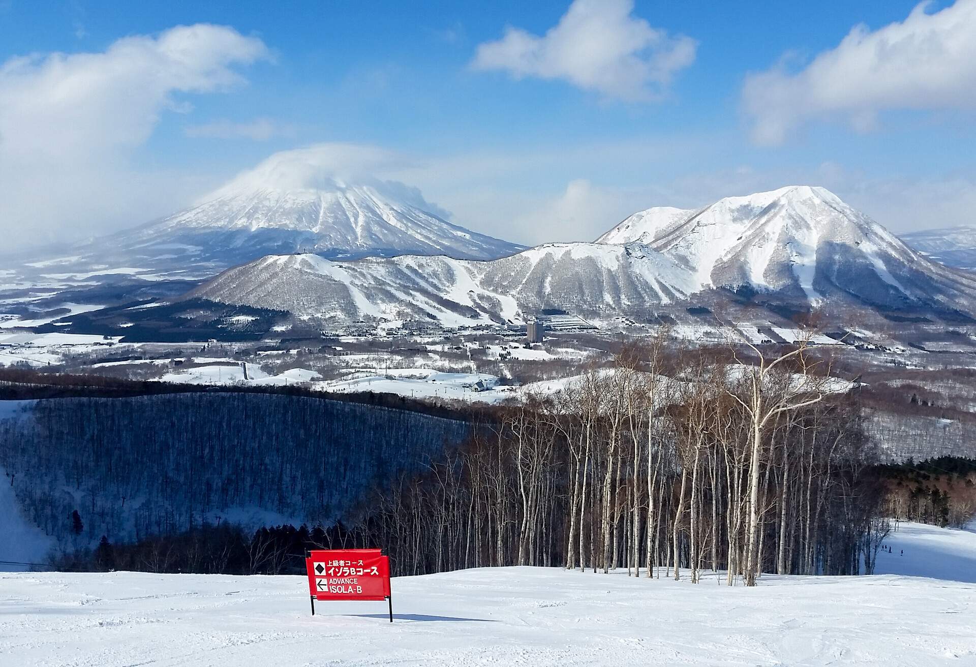 A red caution sign on a hilltop with views of a barren woodland and distant snow-capped mountains.