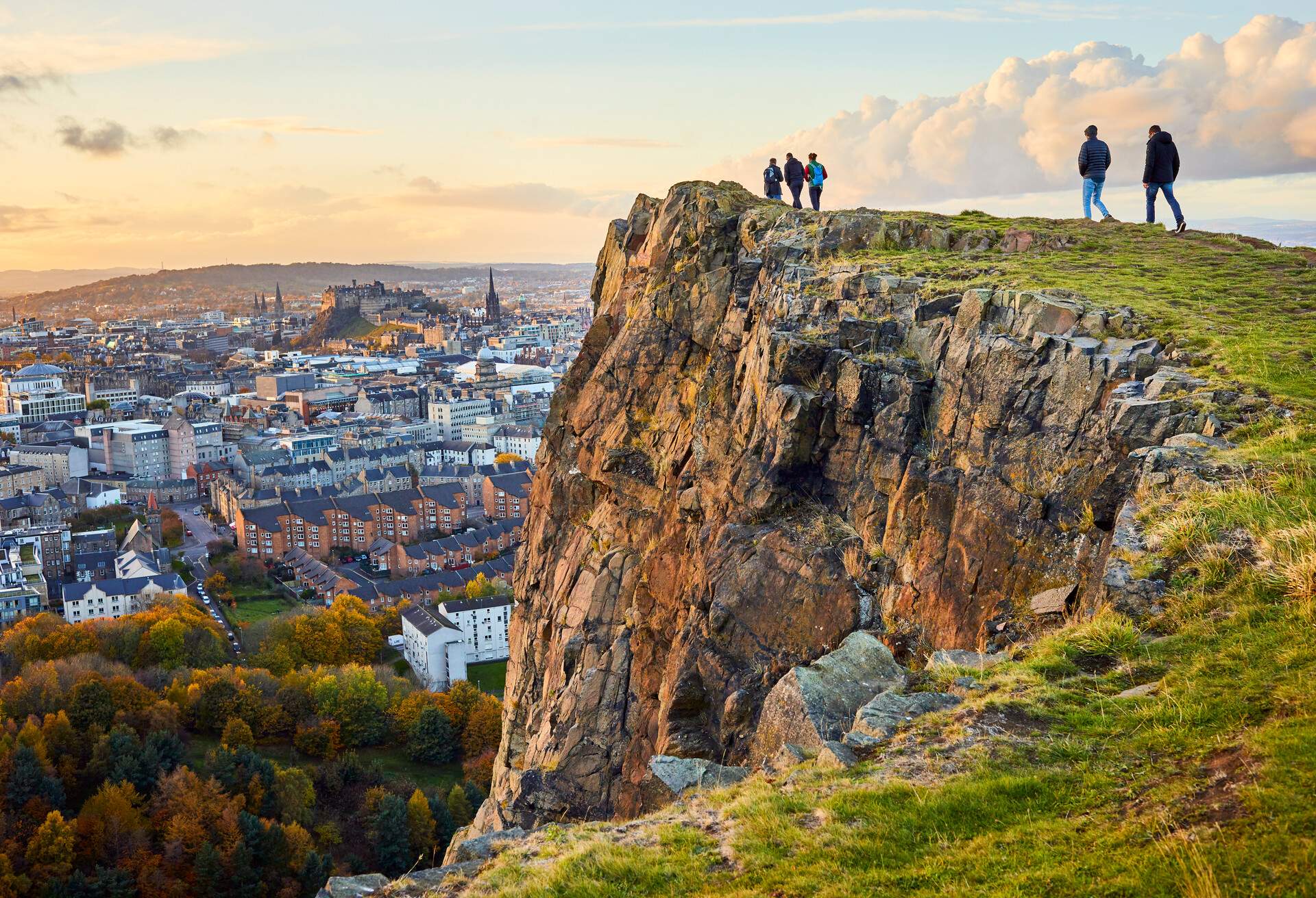 Group of people walking along cliff edge looking at city views. Edinburgh Castle in the distance