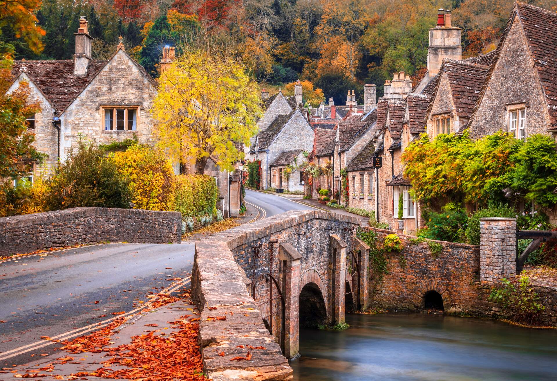 Road bridge leading to a community of Cotswold stone homes with yellow and green autumnal trees.