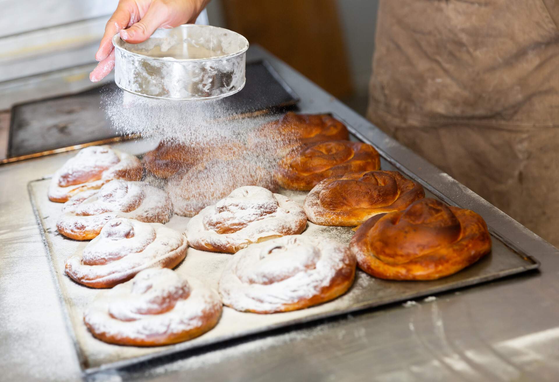 Baker hands dusting freshly baked ensaimadas with powdered sugar in bakery. Baking process of traditional Spanish pastries