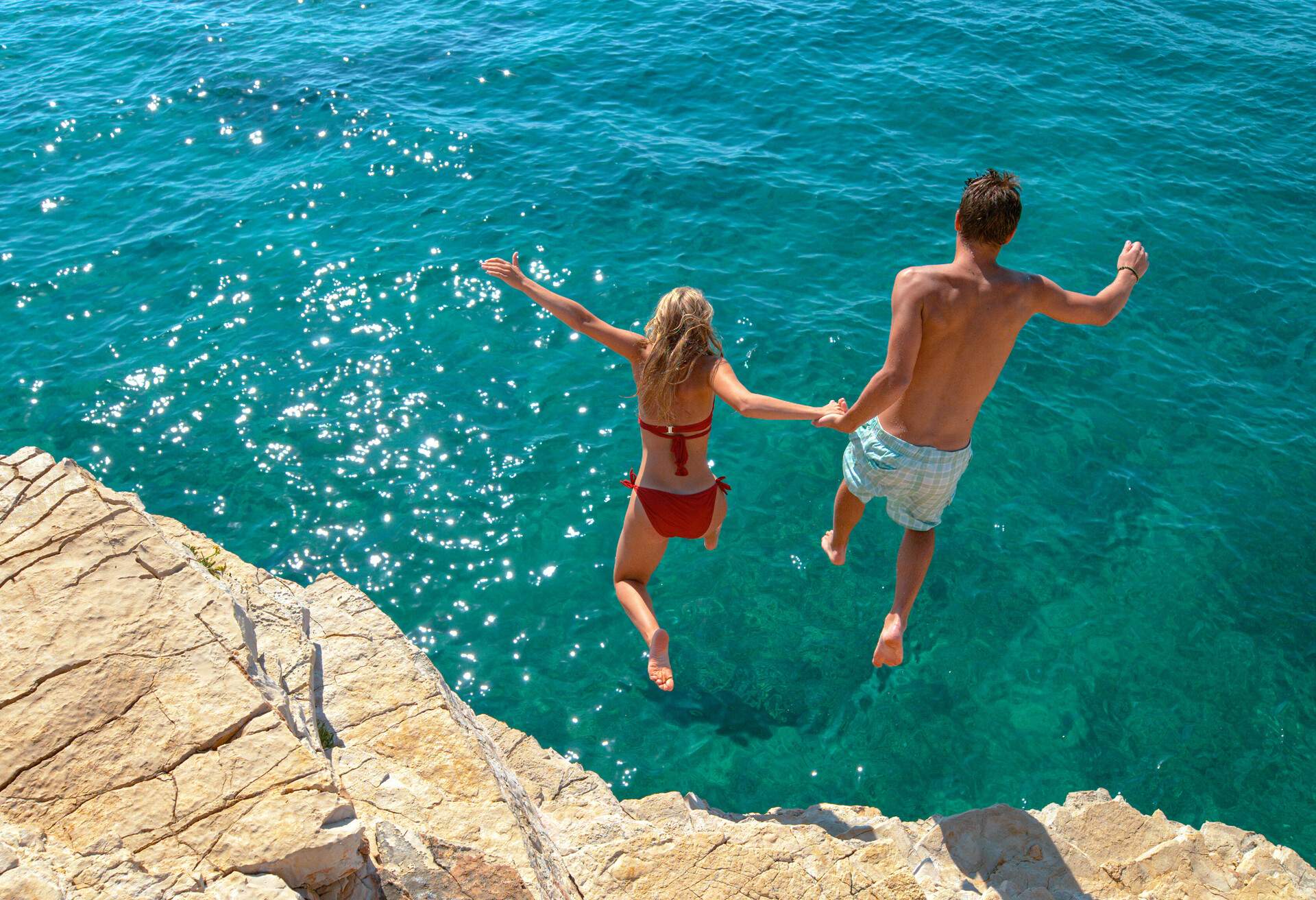 Carefree tourists hold hands while jumping into the refreshing blue sea during a relaxing summer vacation. Active young woman and her boyfriend dive off a high cliff and into the deep blue ocean.