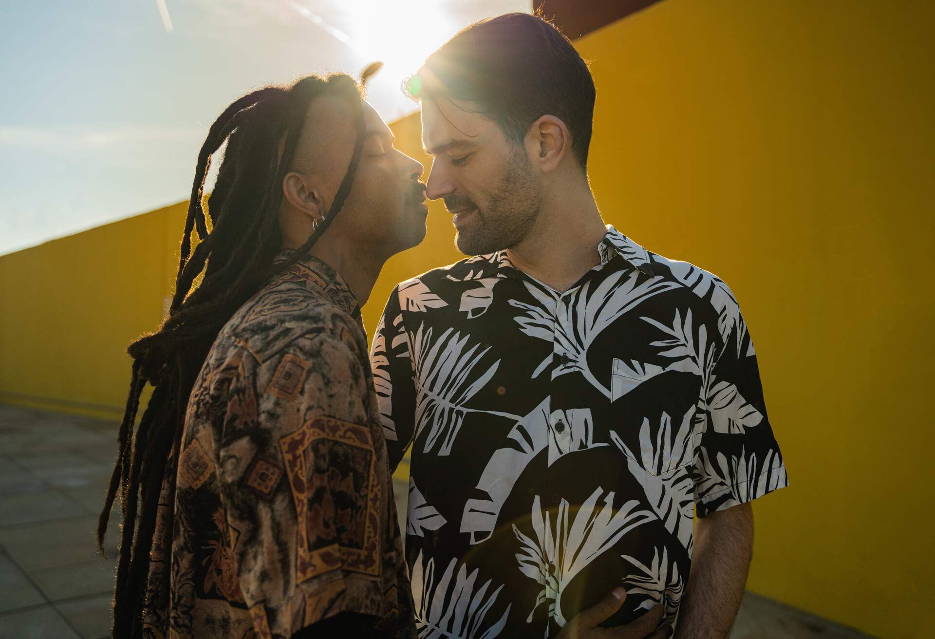 Young gay multiethnic couple looking at each other about to kiss, dressed in flower shirt and are in the city on a yellow background.
