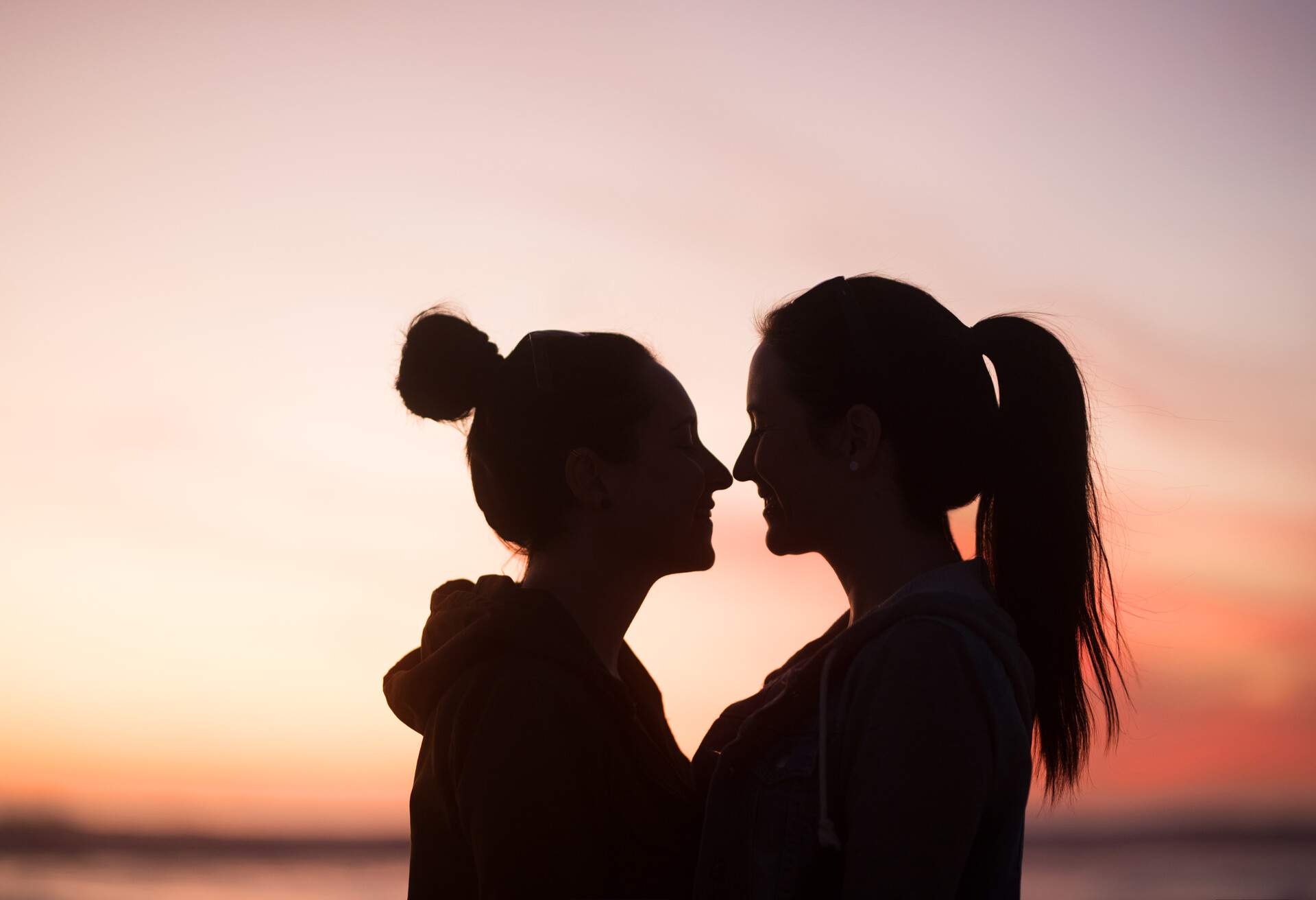 Outline of two young women standing very close to one another looking at each other at sunset in front of the ocean