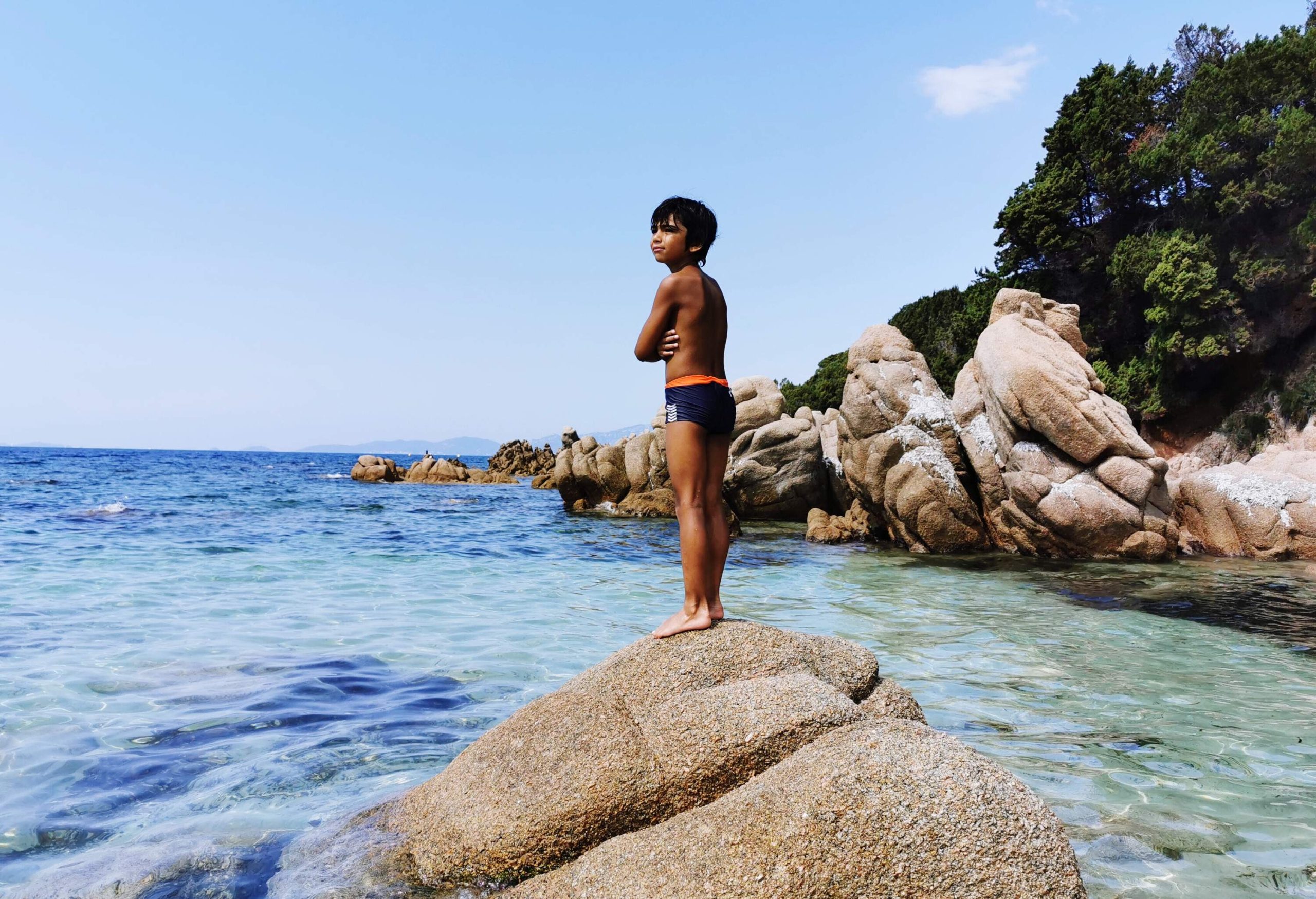A kid in swimwear stands on a giant boulder along a rocky coastline.