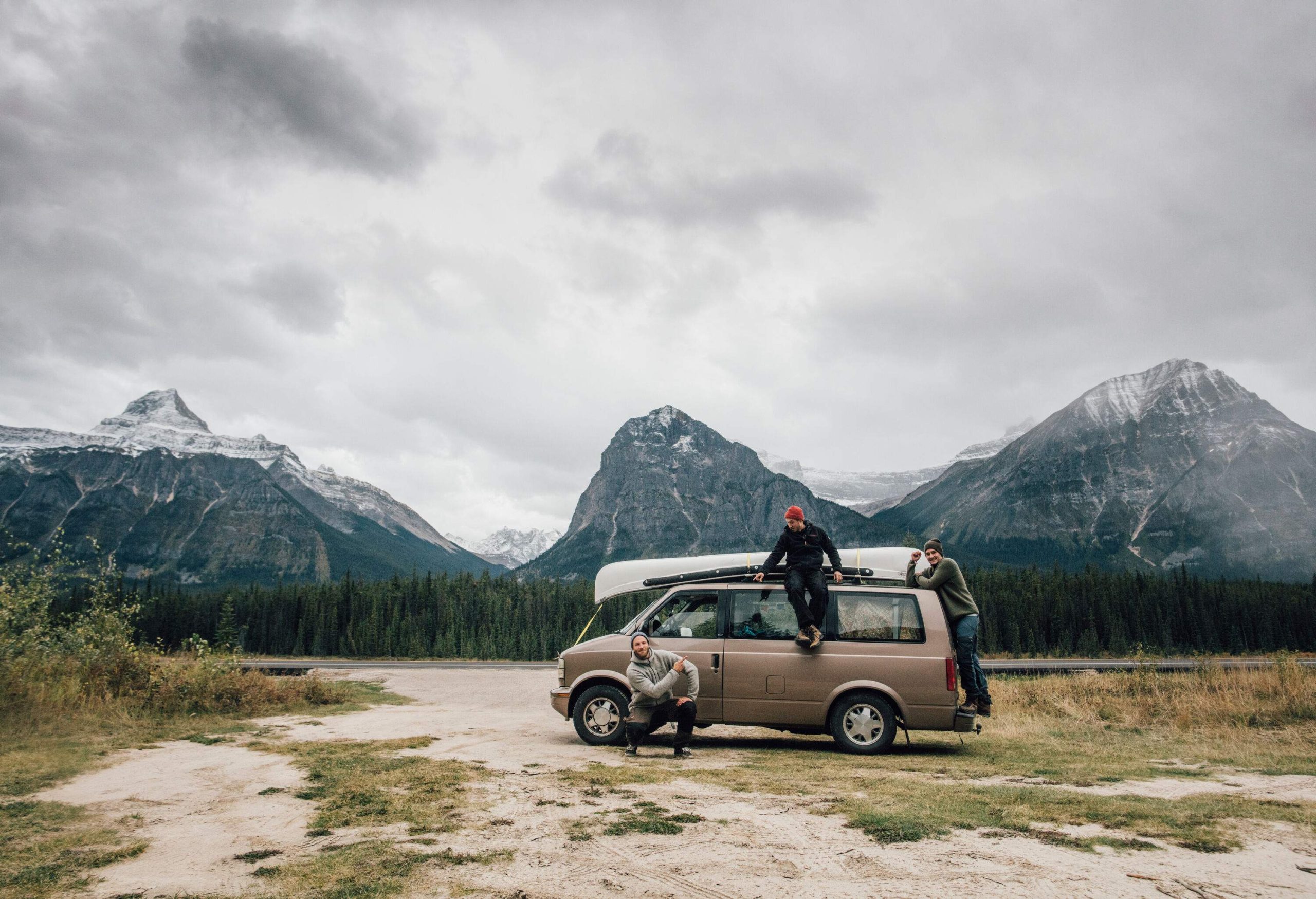 Three friends in winter attire strike playful poses by their trusty van—one perched atop, another riding the back, and the third sitting on the side—all set against a magnificent backdrop of snow-dusted rocky mountains.