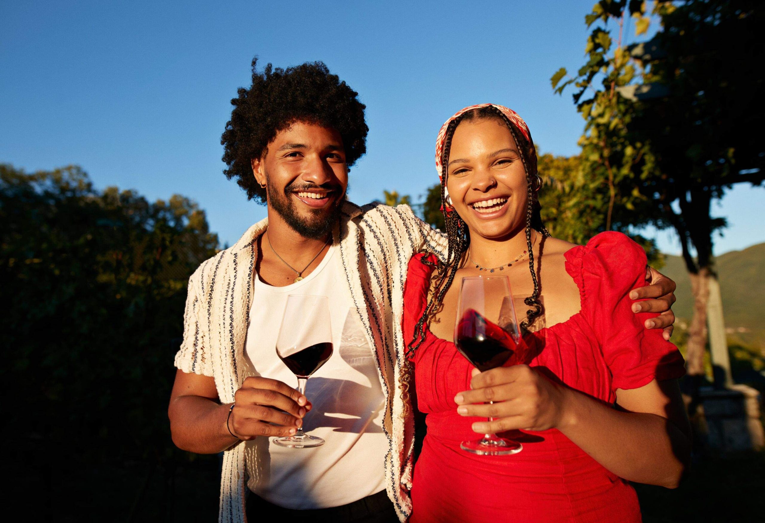 Portrait of happy couple holding wineglasses standing with arm around each other in vineyard during sunset
