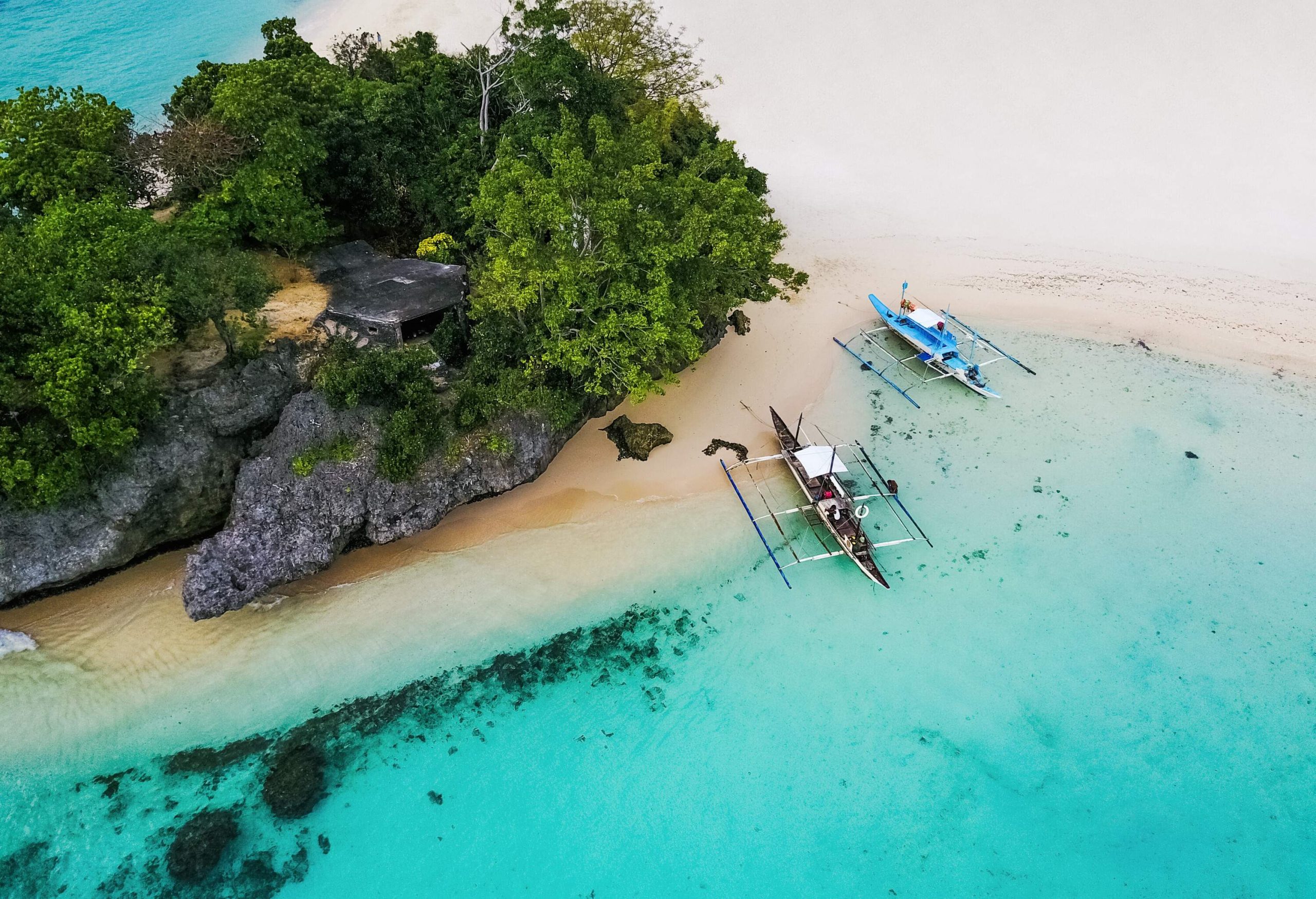 A rock cliff with lush vegetation along a white beach with anchored fishing boats.