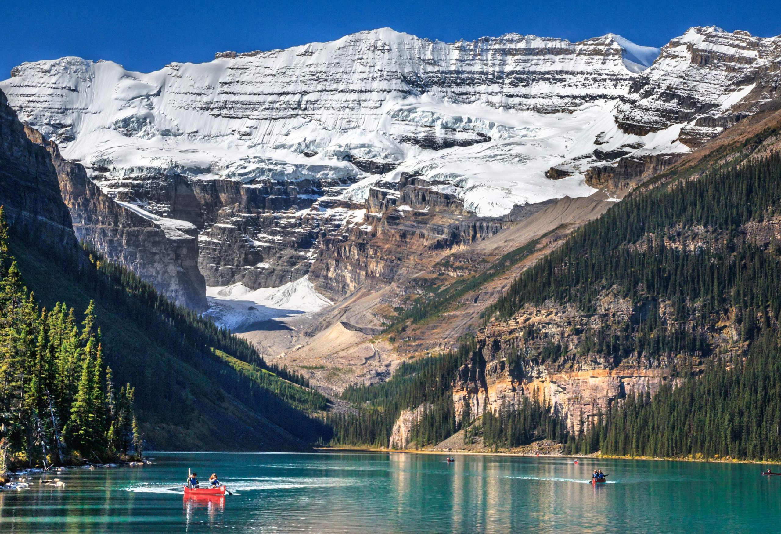 People riding a canoe on a lake between mountains covered in coniferous trees and snow.