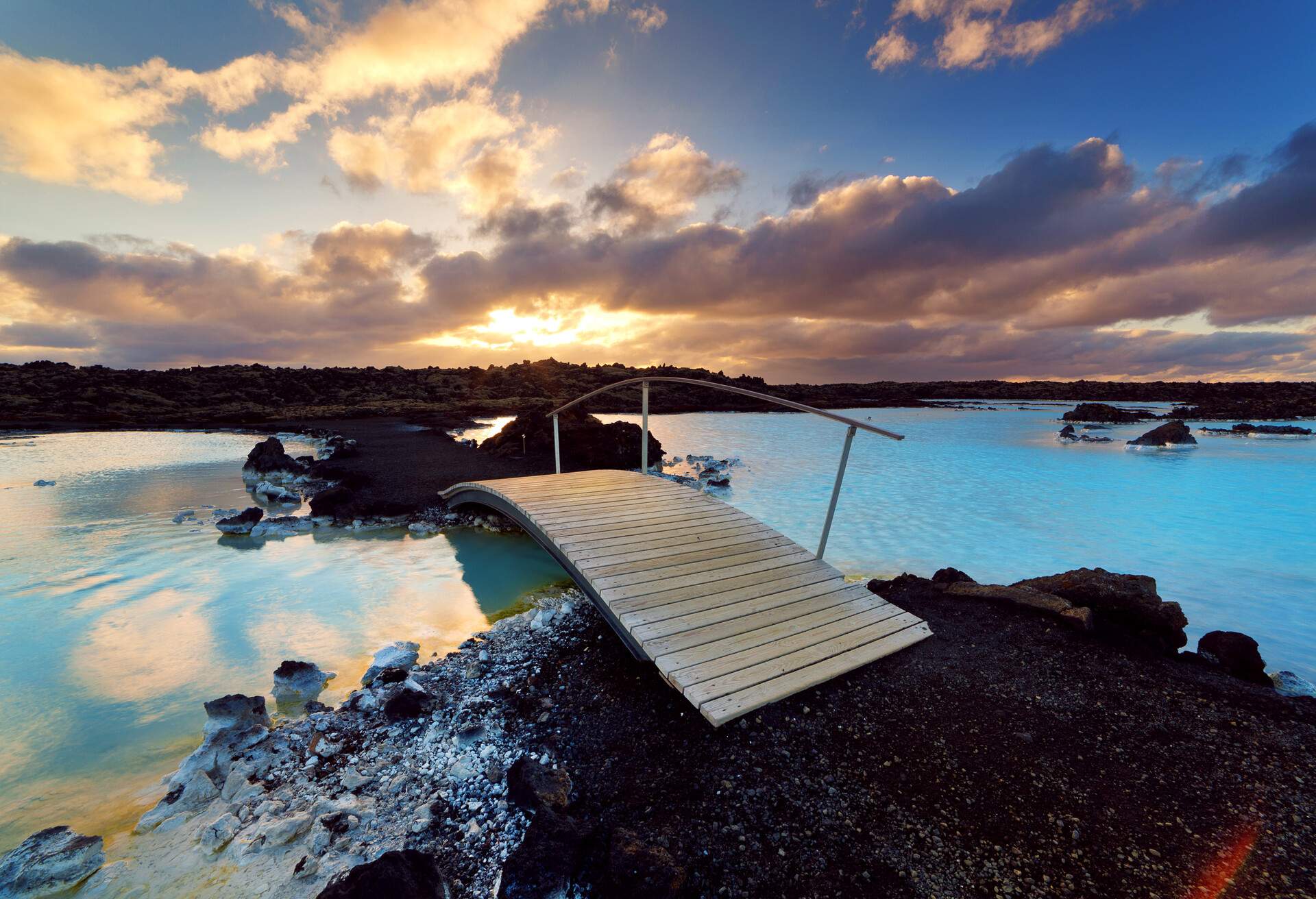 Sunset over railed footbridge surrounded by volcanic landscape at Blue Lagoon, Iceland, with aquamarine water reflecting colors of spring sunset.