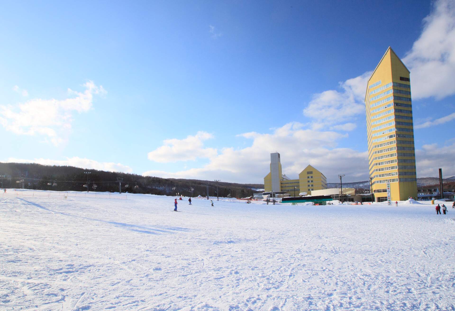 A yellow structure with glass windows adjacent to a snowfield with skiers.