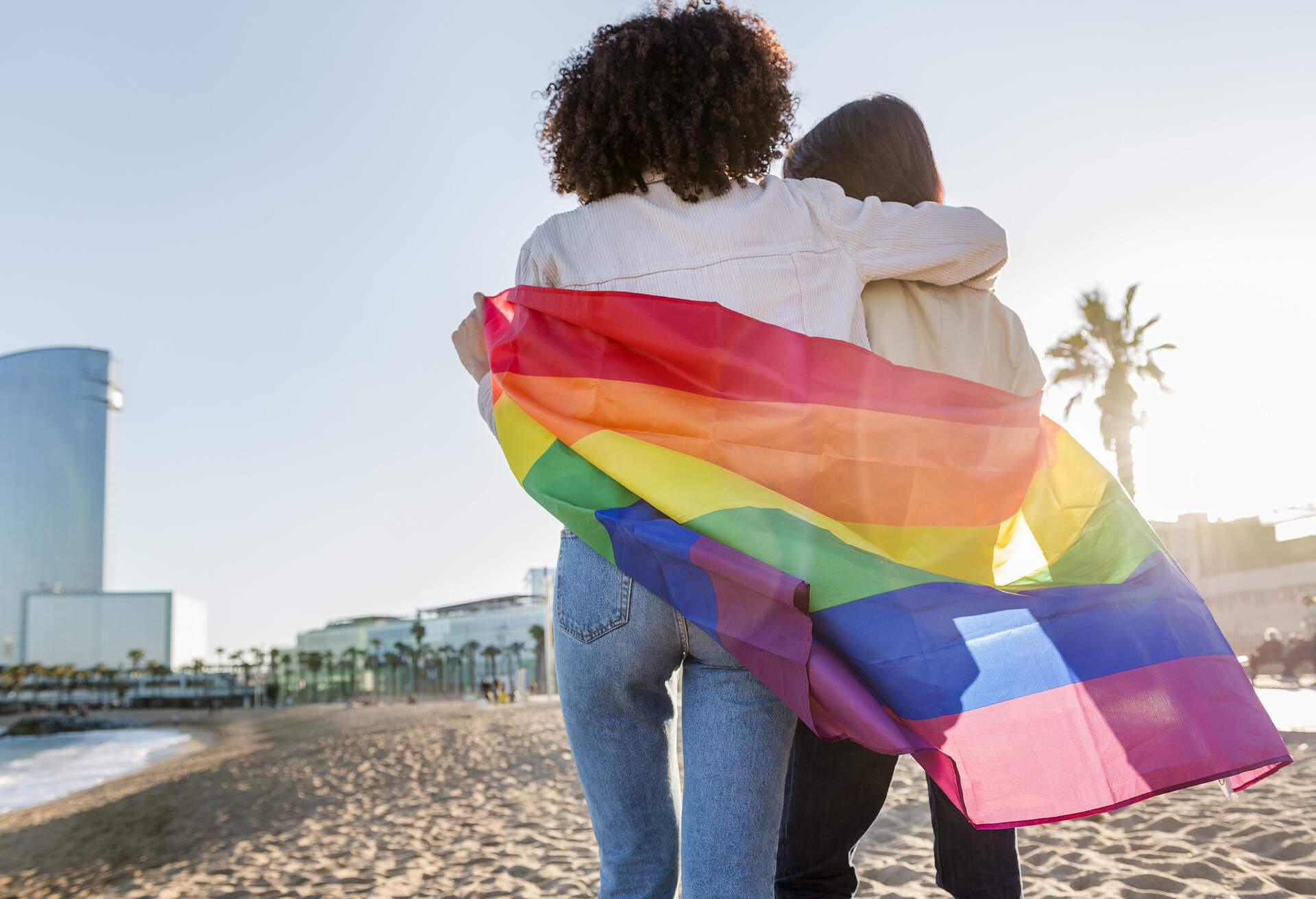 Two people shrouded in a Pride flag on the beach.