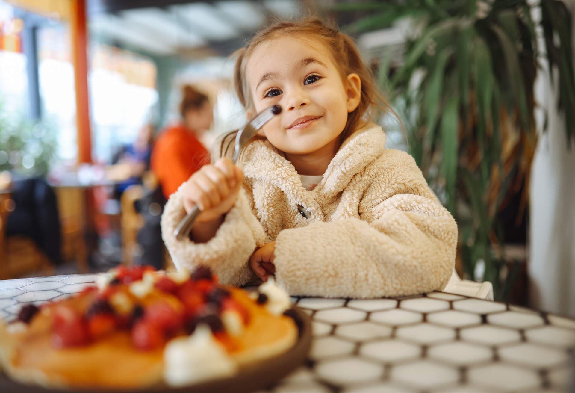 Little girl having crepes for breakfast. The crepes are sprinkled with fruits - blueberries and raspberries. Sweet toddler birthday boy, eating belgian waffle with raspberries and chocolate at cafe