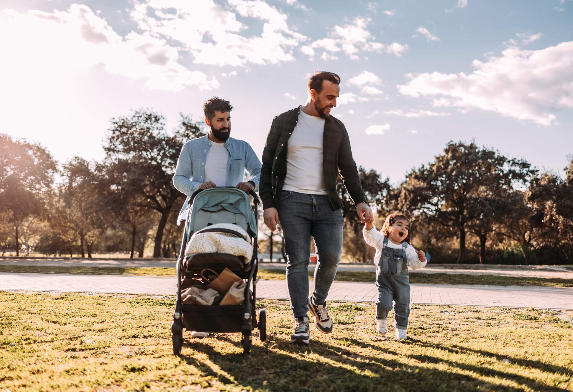 Gay fathers and their young daughter walking in the park. One of them is pushing the pram and the other is holding the small child's hand.