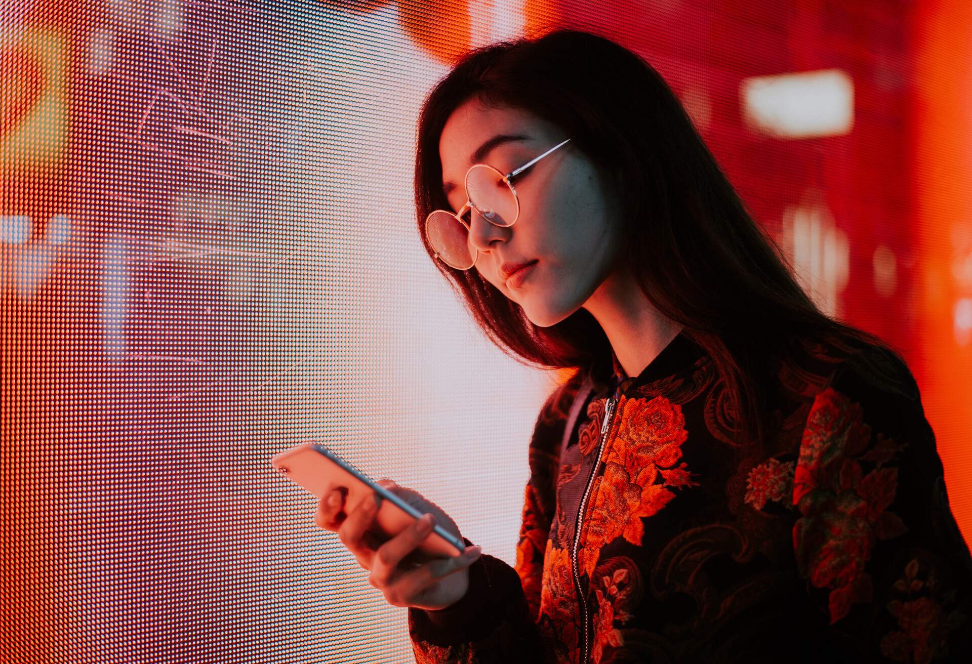 Woman with dark hair, glasses, and a floral jacket holds her phone and glanced at it.