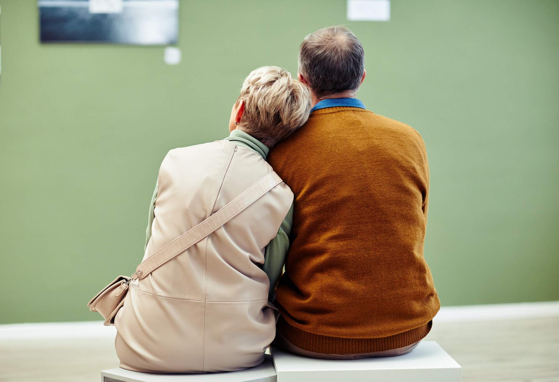 Rear view of unrecognizable mature man and woman in love sitting on stools in modern art gallery looking at artworks