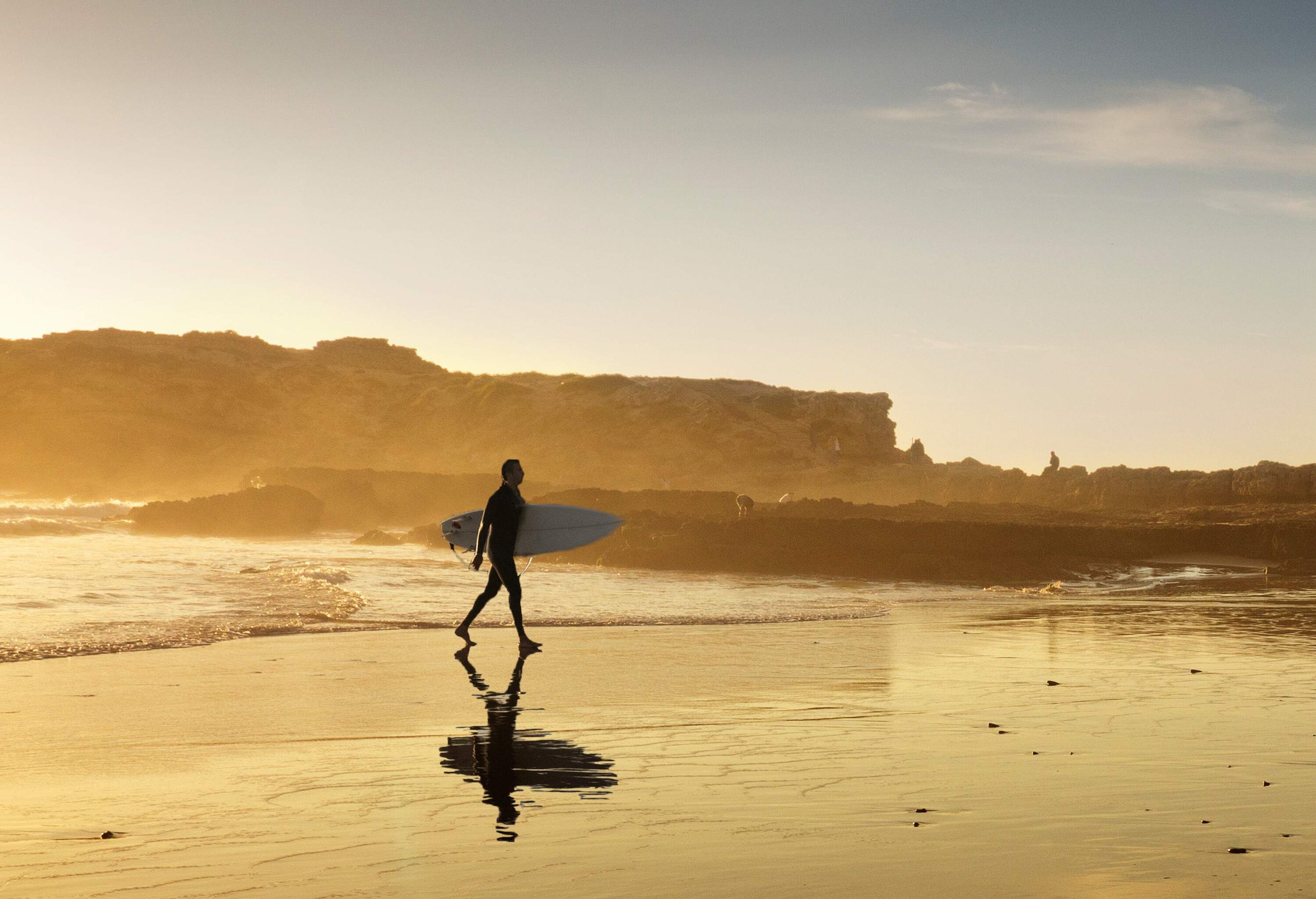 A man carrying a surfboard as he walks across the beach towards the coast.