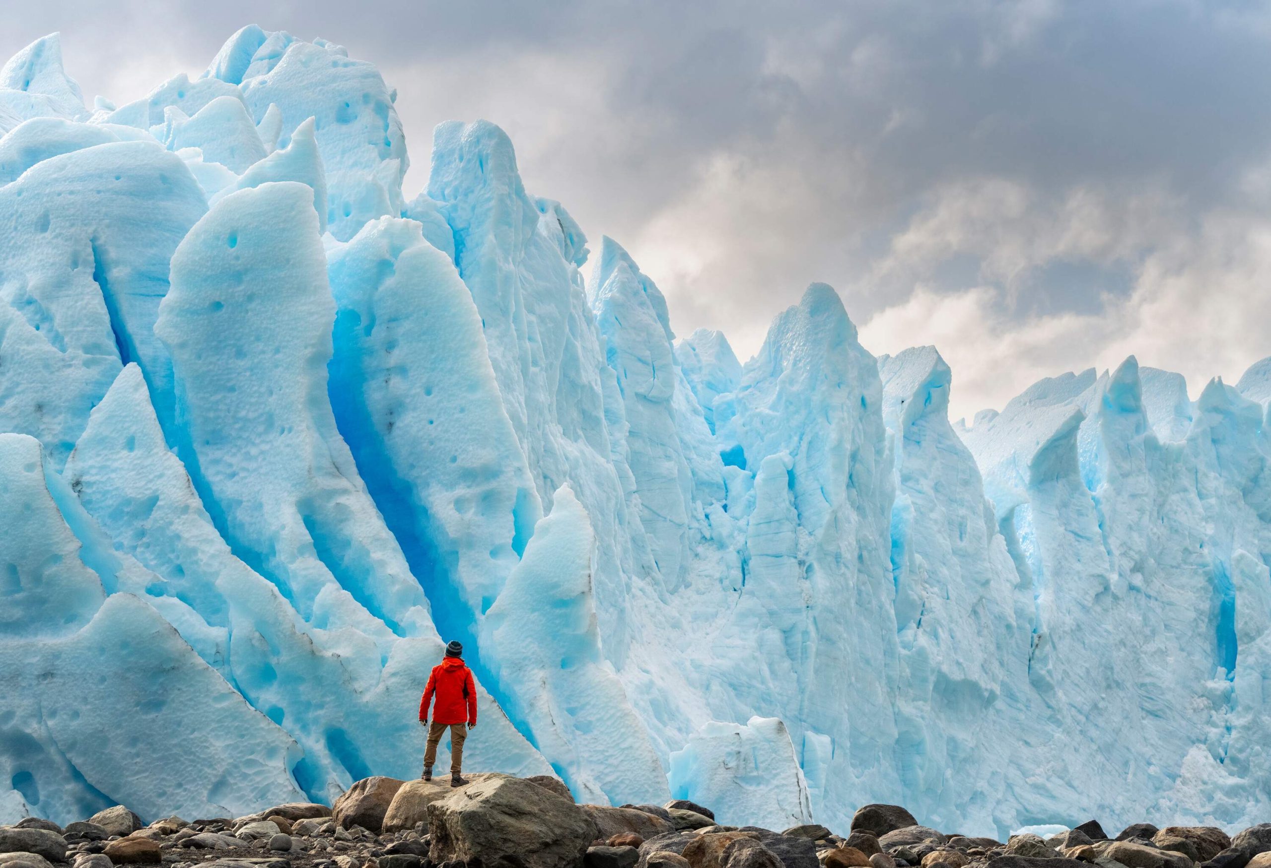 Man in an orange jacket stands on a boulder and watches the glacier's soaring ice formations.