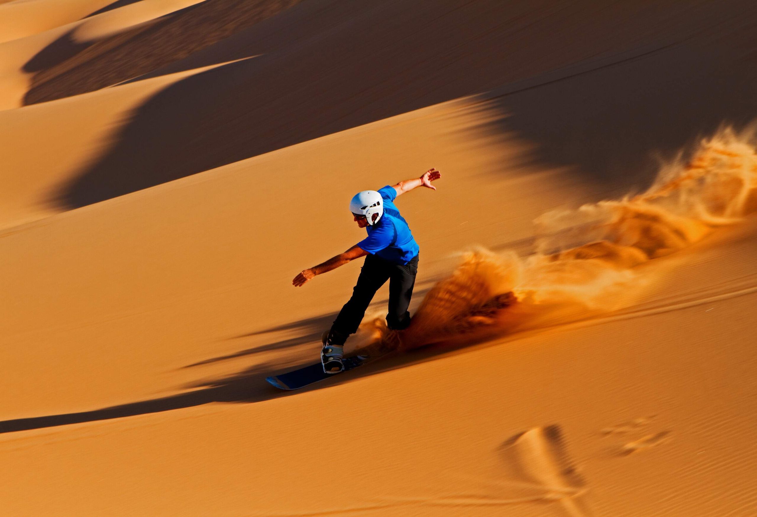 A man in a blue shirt and a white helmet sandboarding, leaving a trail of dust behind him.
