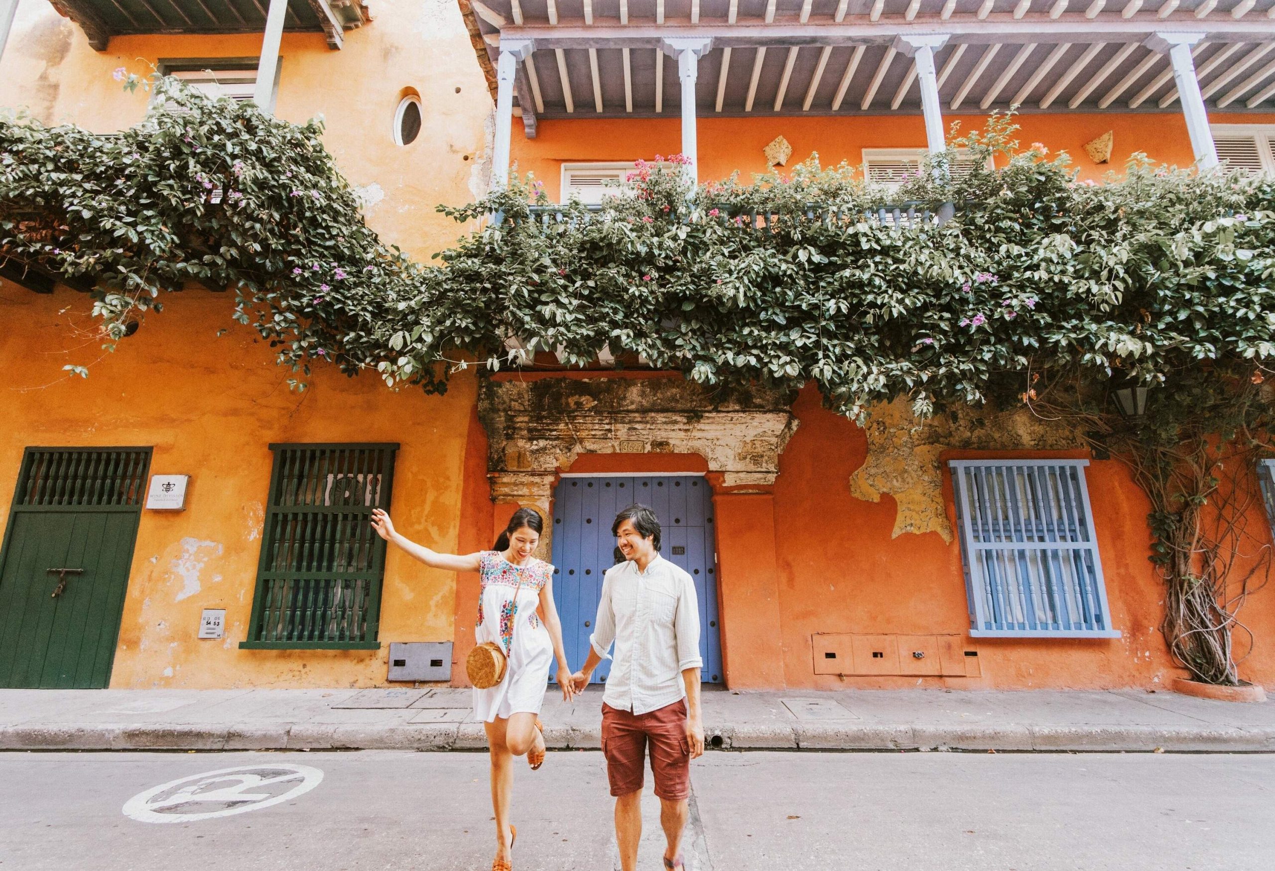 A couple in casual clothes holds hands as they stand in front of an orange house with green vines all over its balcony.