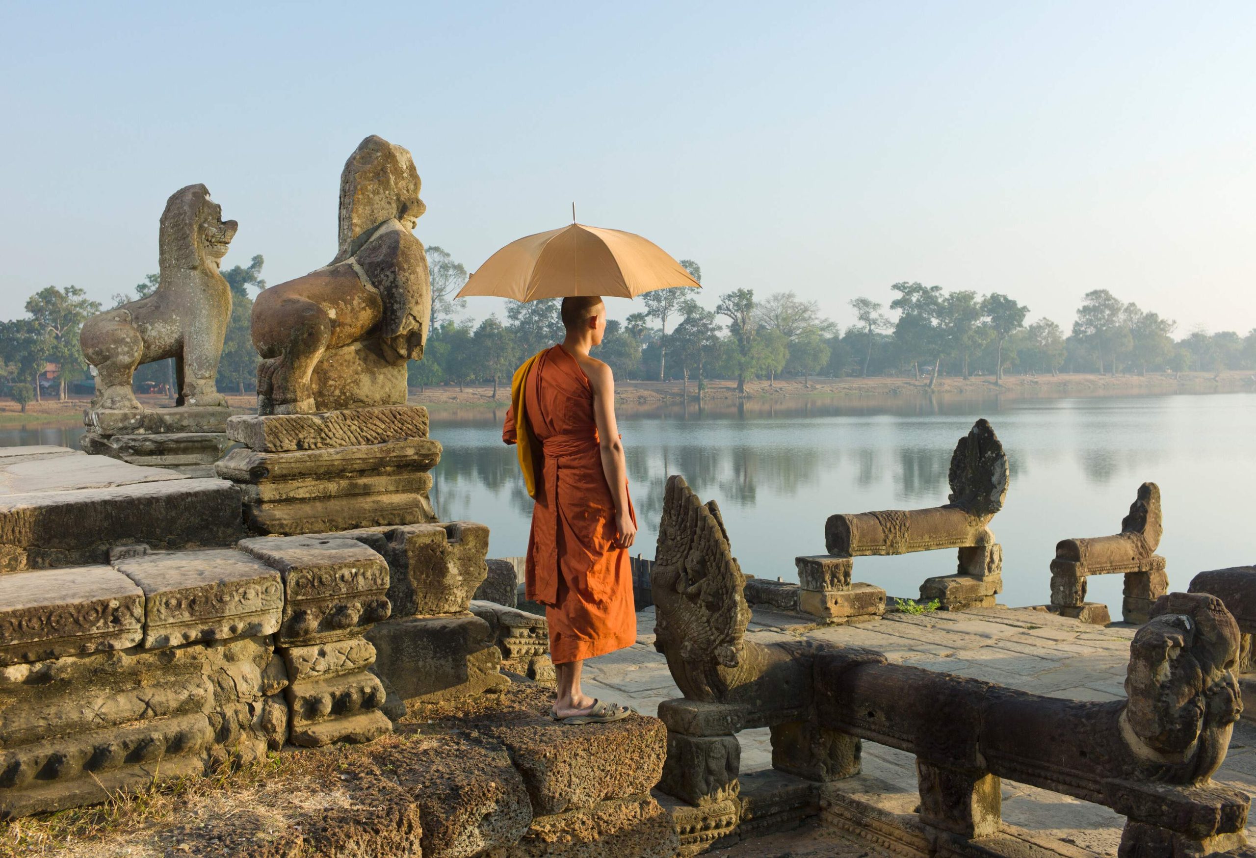 A monk stands next to the stone sculptures of the temple, gazing at the lake while holding an umbrella.