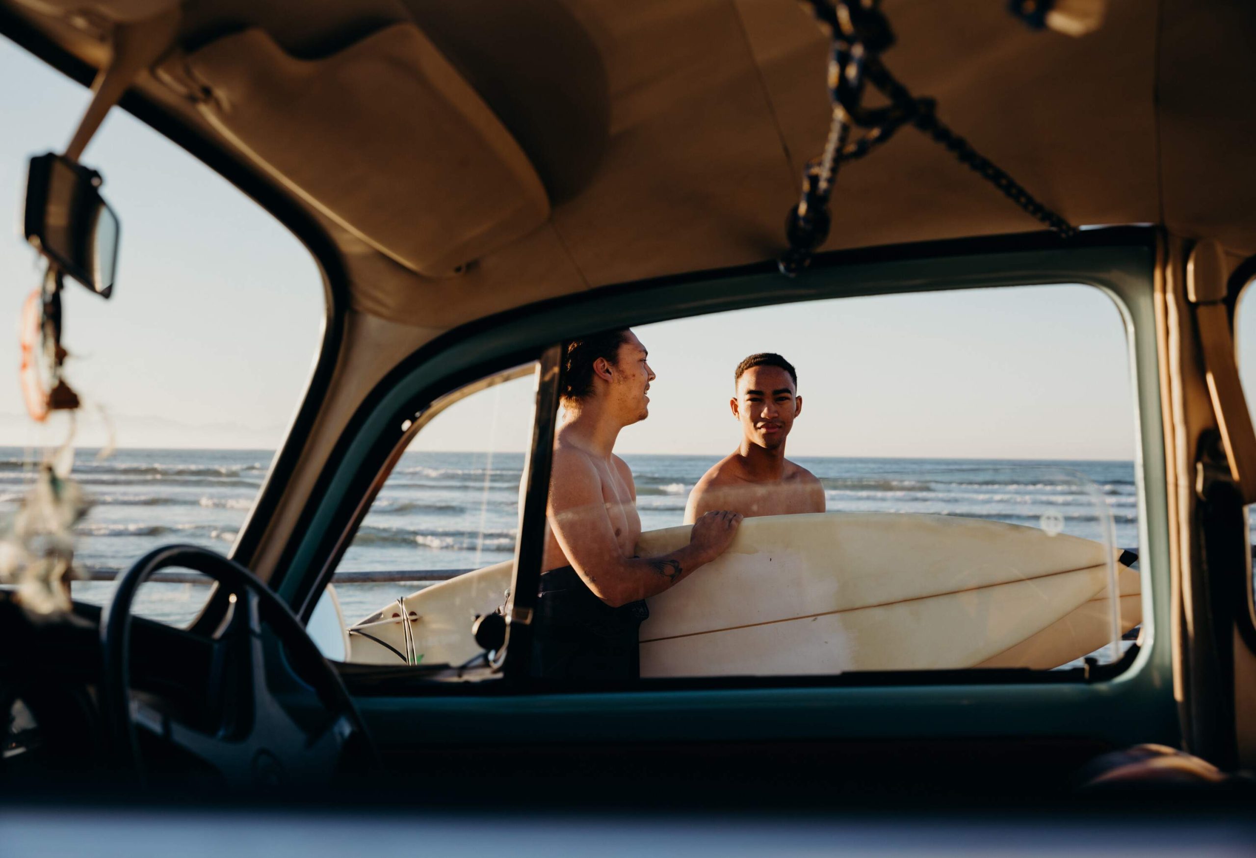 Two men walk on a beach with their surfboards as seen from the interior of a car.