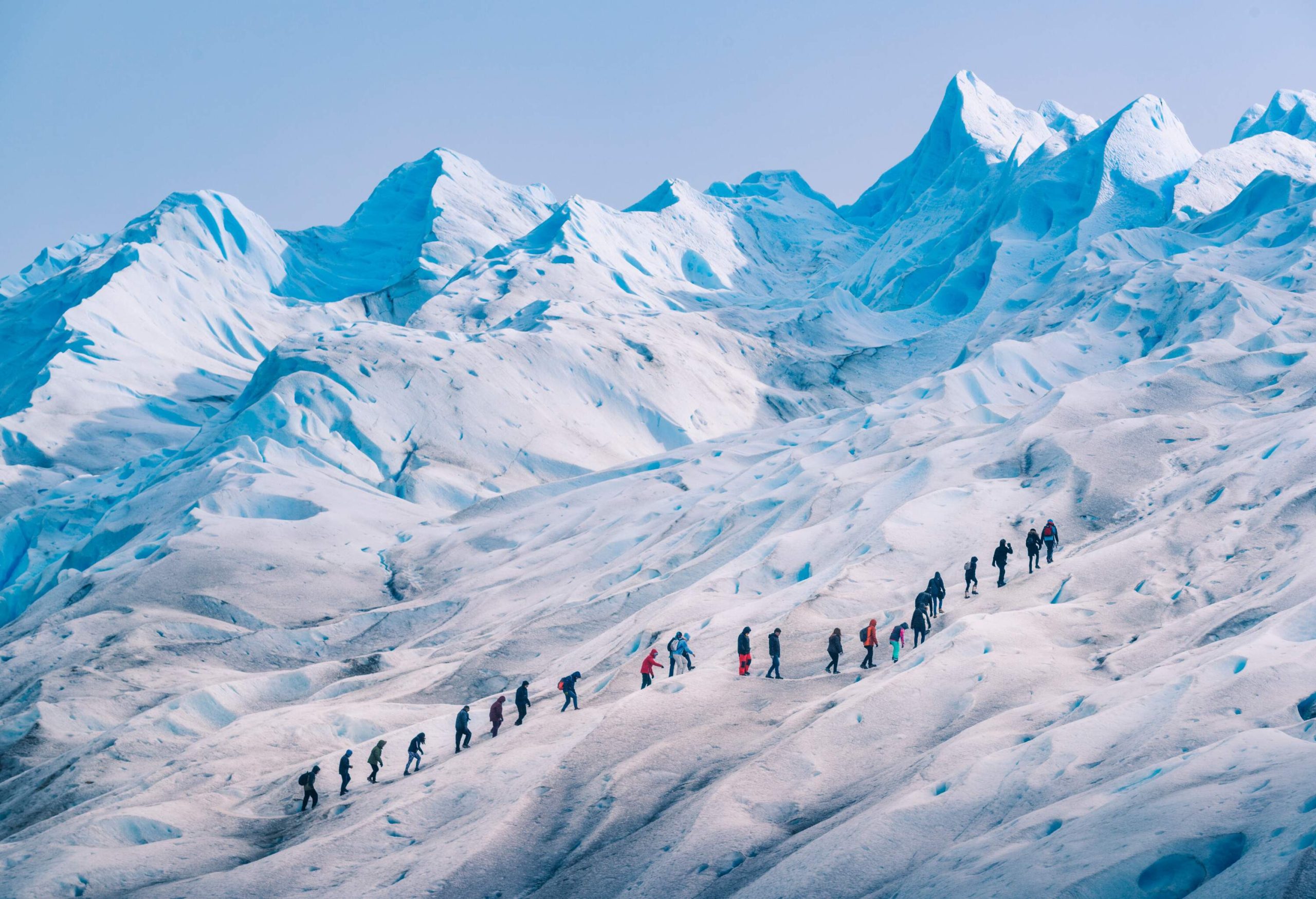 A row of hikers walking on a glacier with jagged peaks.