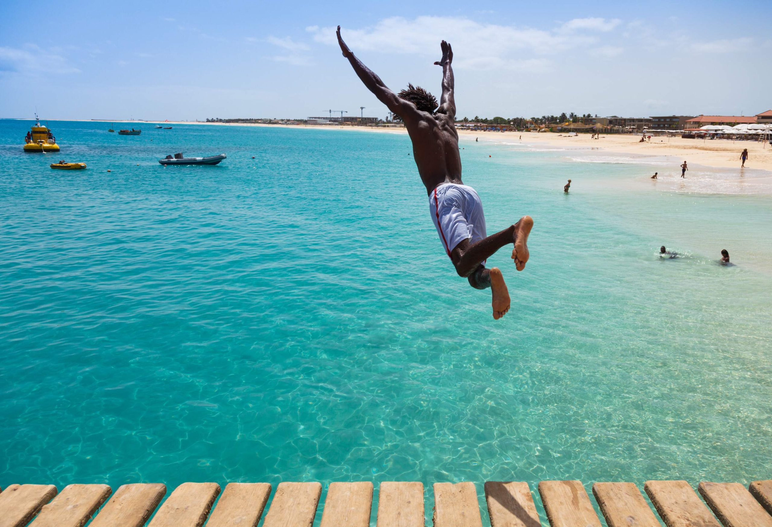 A man in boardshorts jumps into a beach's turquoise waters from a wooden pier.