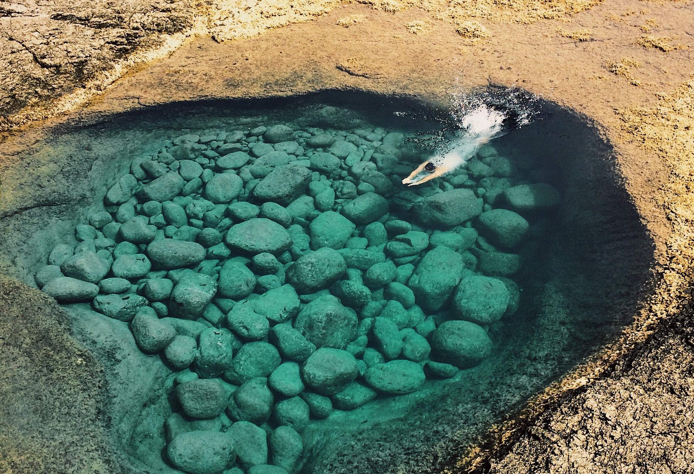 A man dives into a natural rock pool with crystalline green colour water.