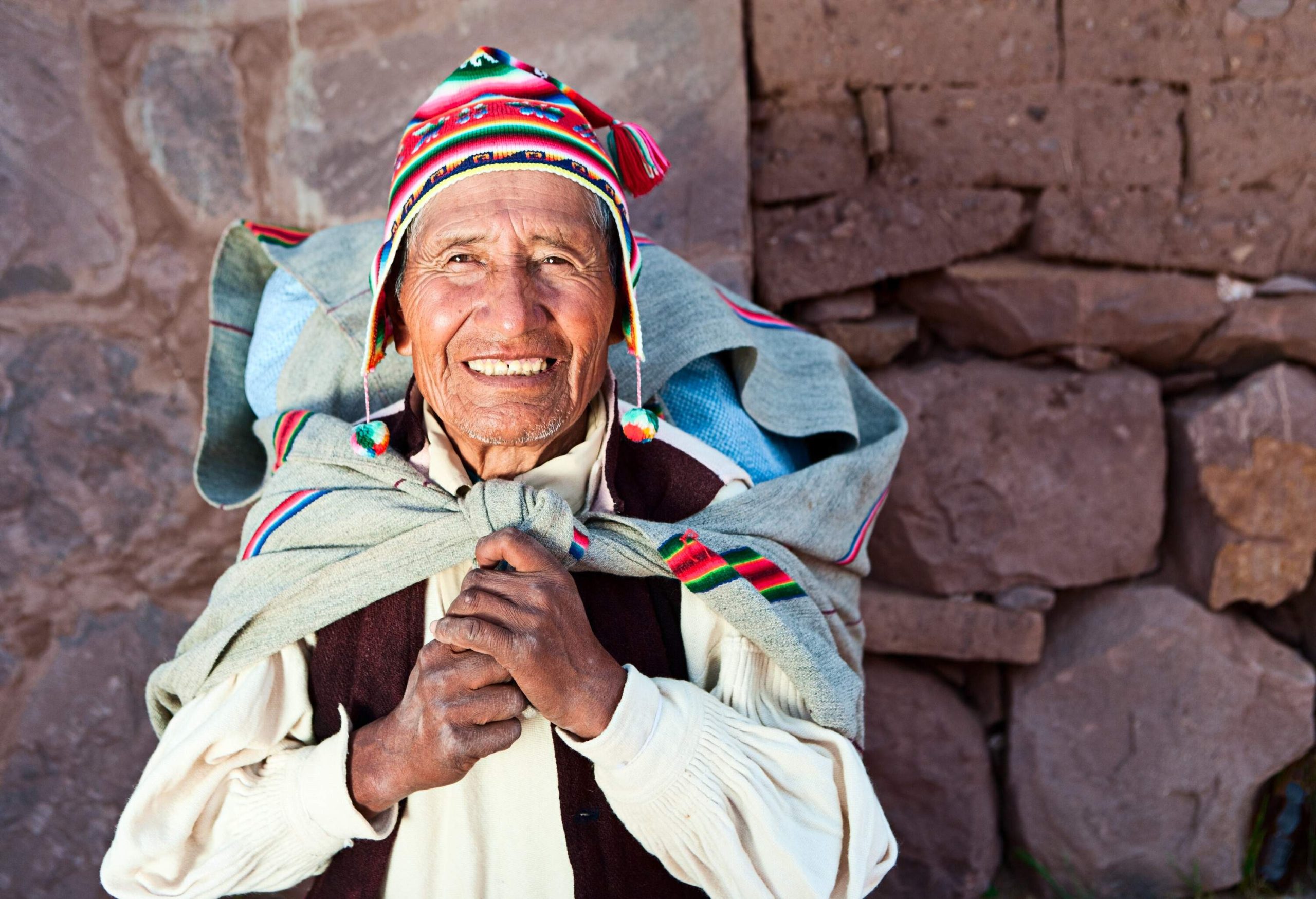 A cheerful old man wearing a vibrant knitted cap and jacket, along with a sack bag, beams with a warm smile.