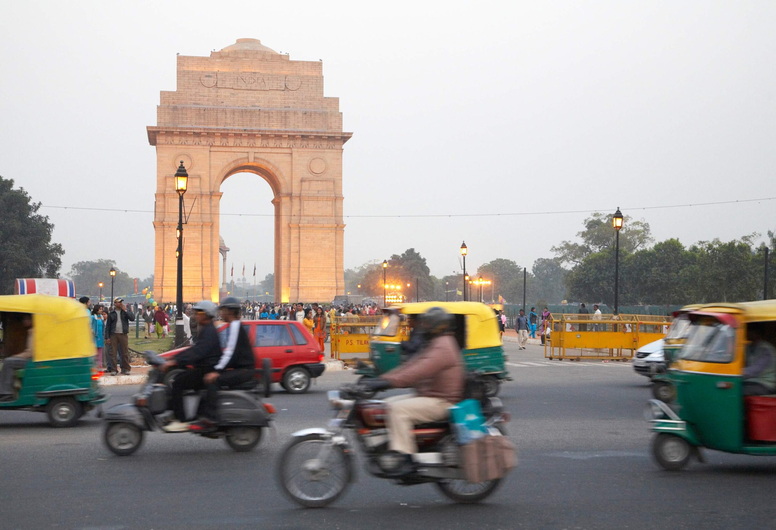 Motorcycles and rickshaws travelling down the street, with the India Gate in the backdrop.