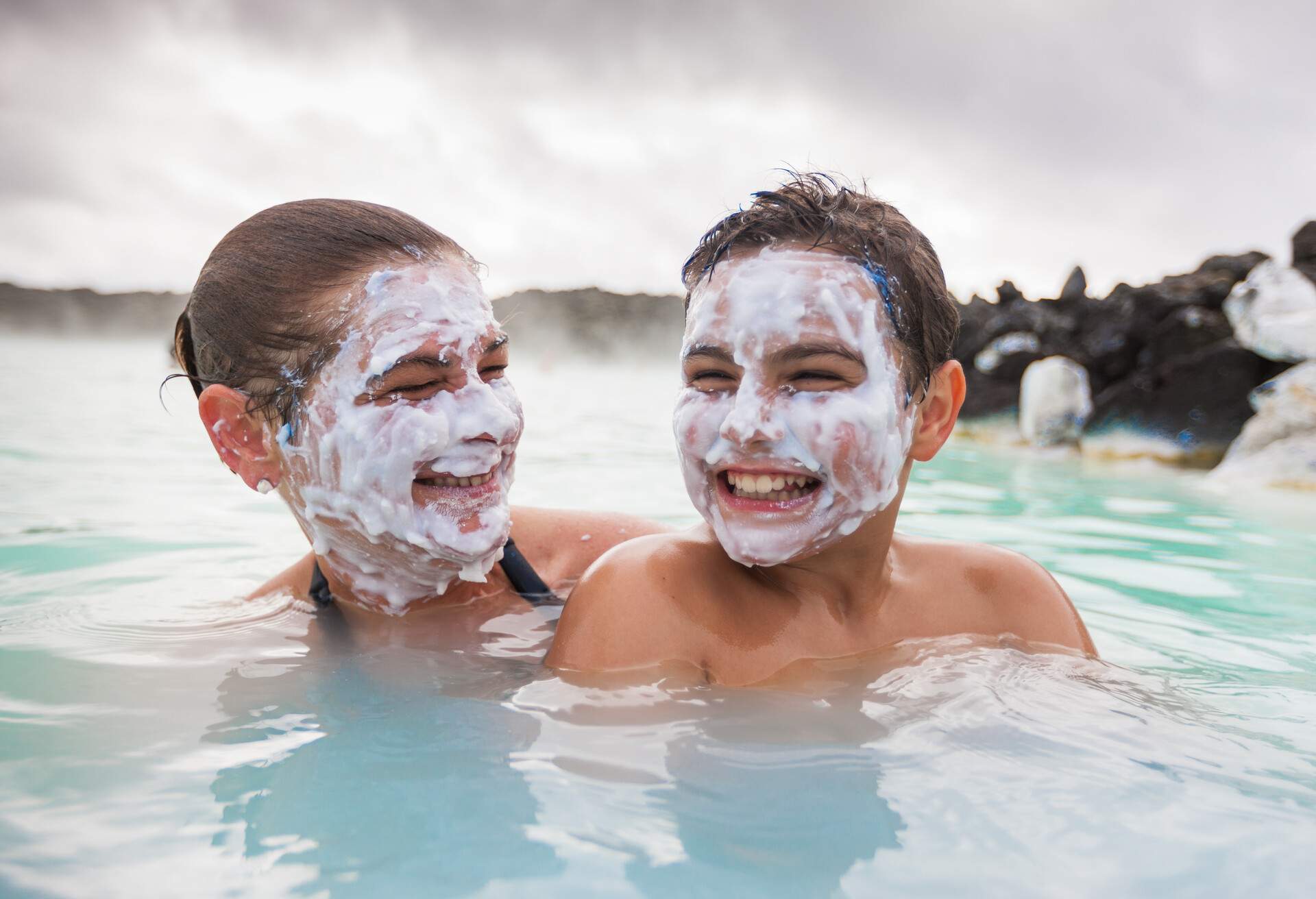 Mother and son having fun together, joking, smiling with natural sulphur face masks in Icelandic Natural Wellness Lagoon. Kevlavic, Iceland.