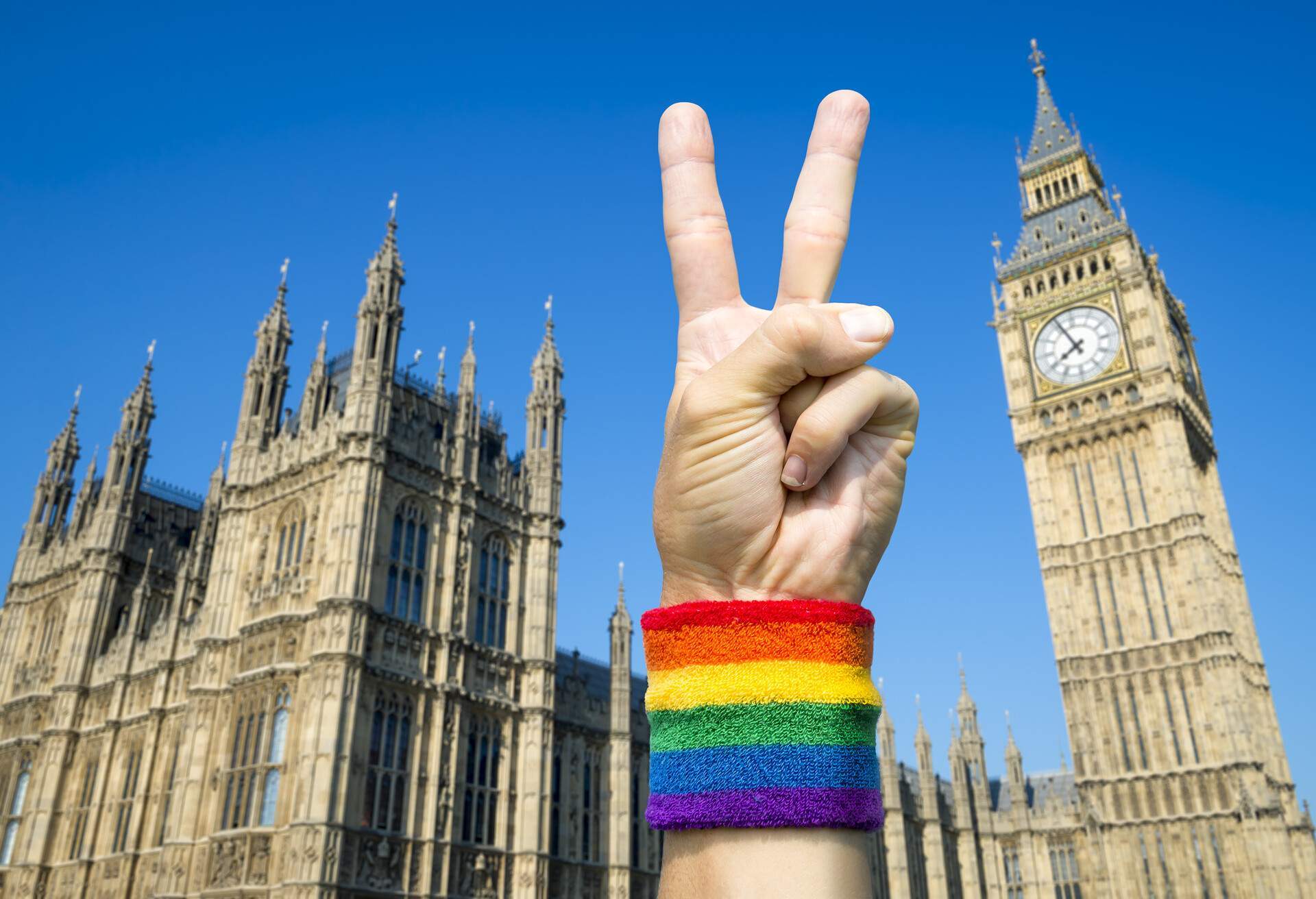 Hand wearing gay pride rainbow flag wristband holding up a victory/peace sign gesture in front of the Houses of Parliament at Westminster, London, UK