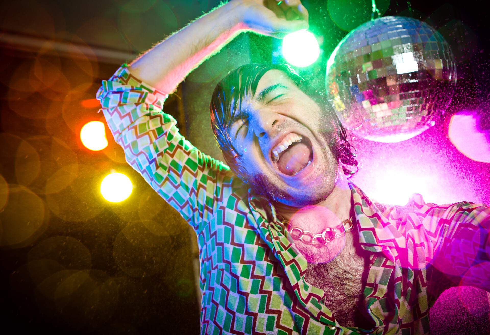 A man in a retro shirt and a chain necklace dancing happily next to a disco ball.